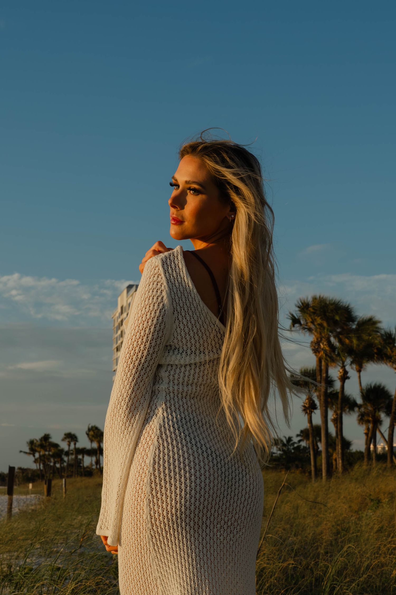Blonde woman in white knit dress standing on a sandy beach at golden hour with palm trees behind her.