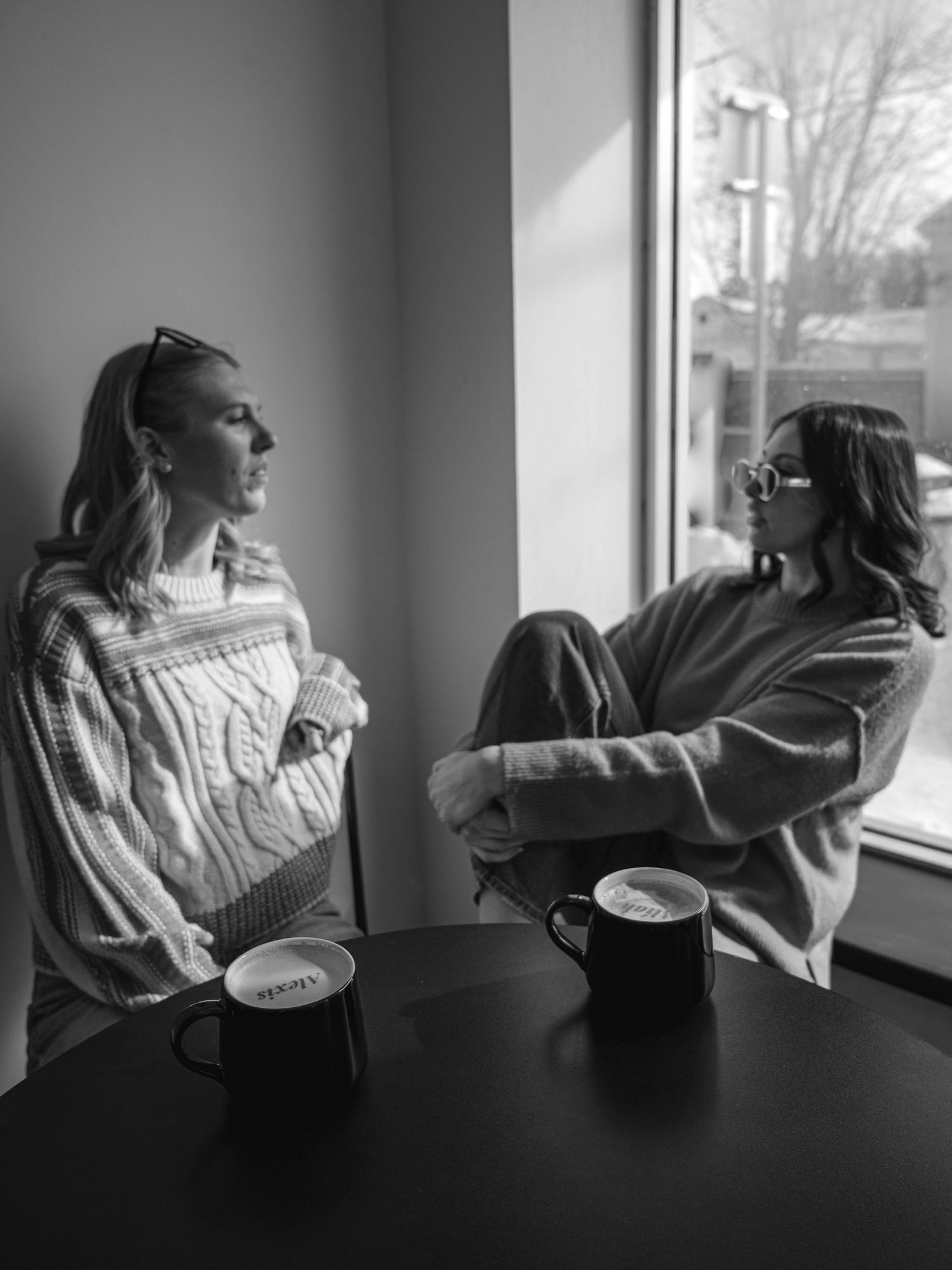Black and white photo of two women conversing over mugs by a sunlit window.