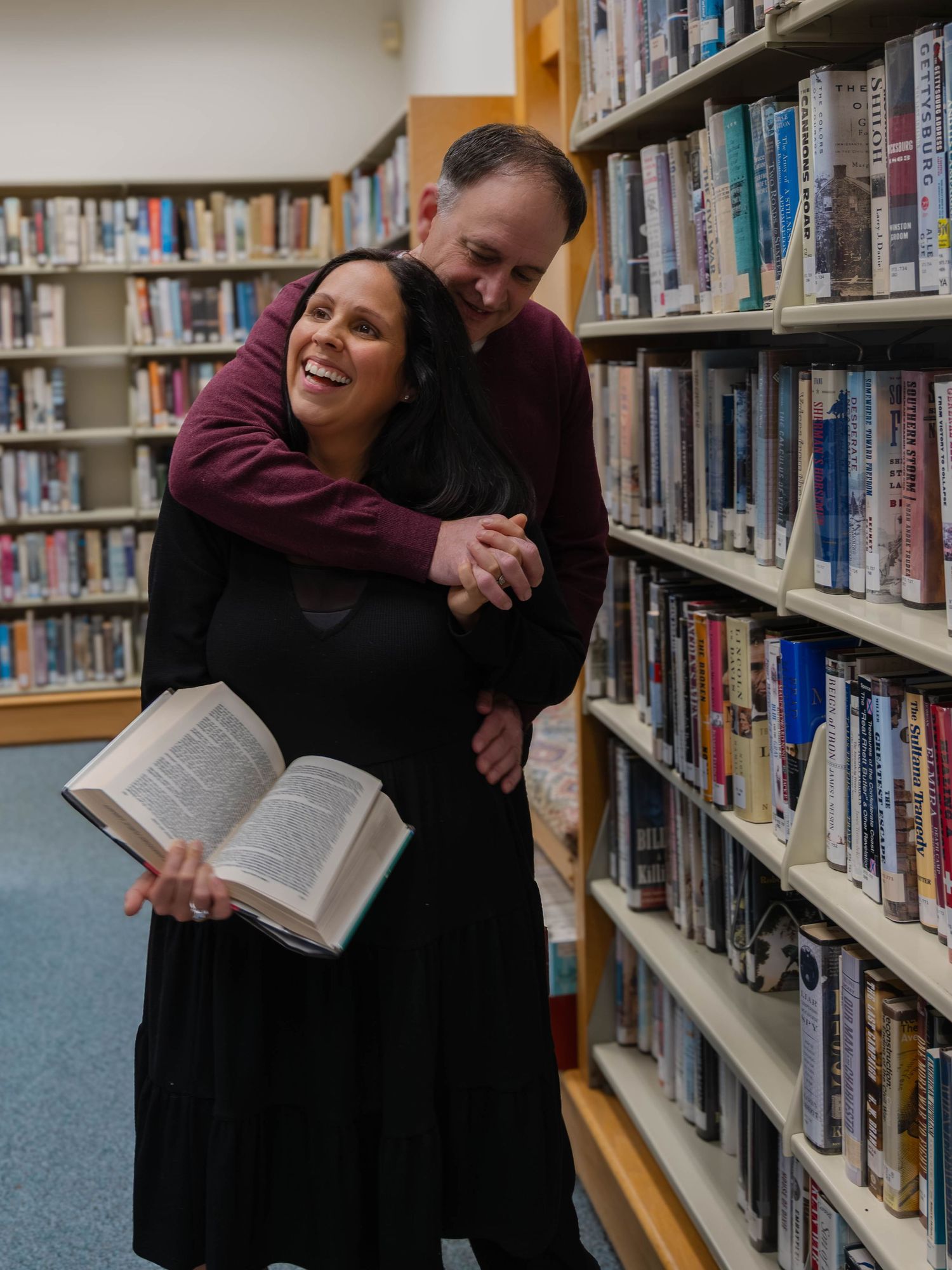 Laughing woman embraced from behind by man in library, holding an open book near bookshelves.