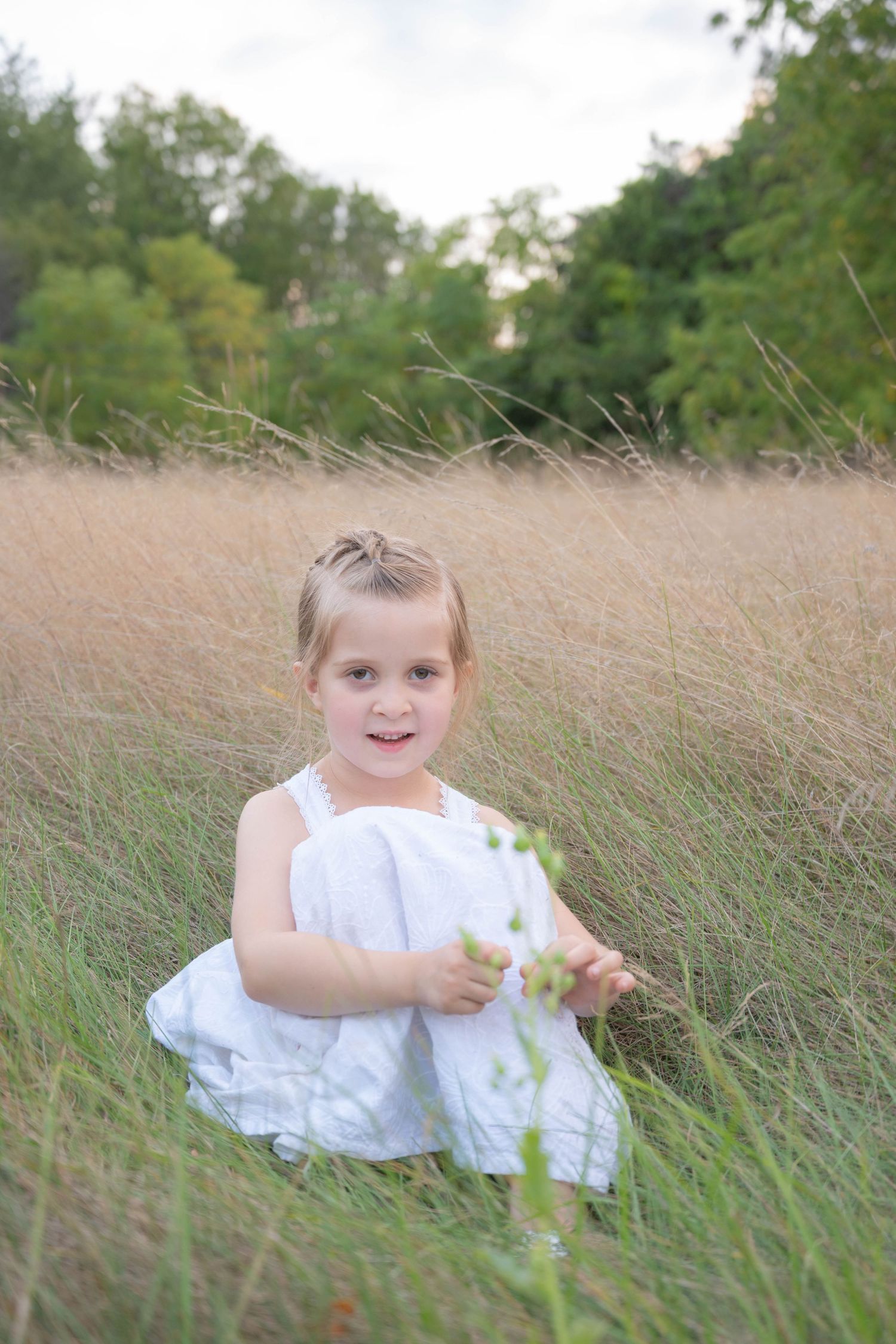 Cheerful little girl in white dress sitting in lush green and golden grass field with trees in the background.