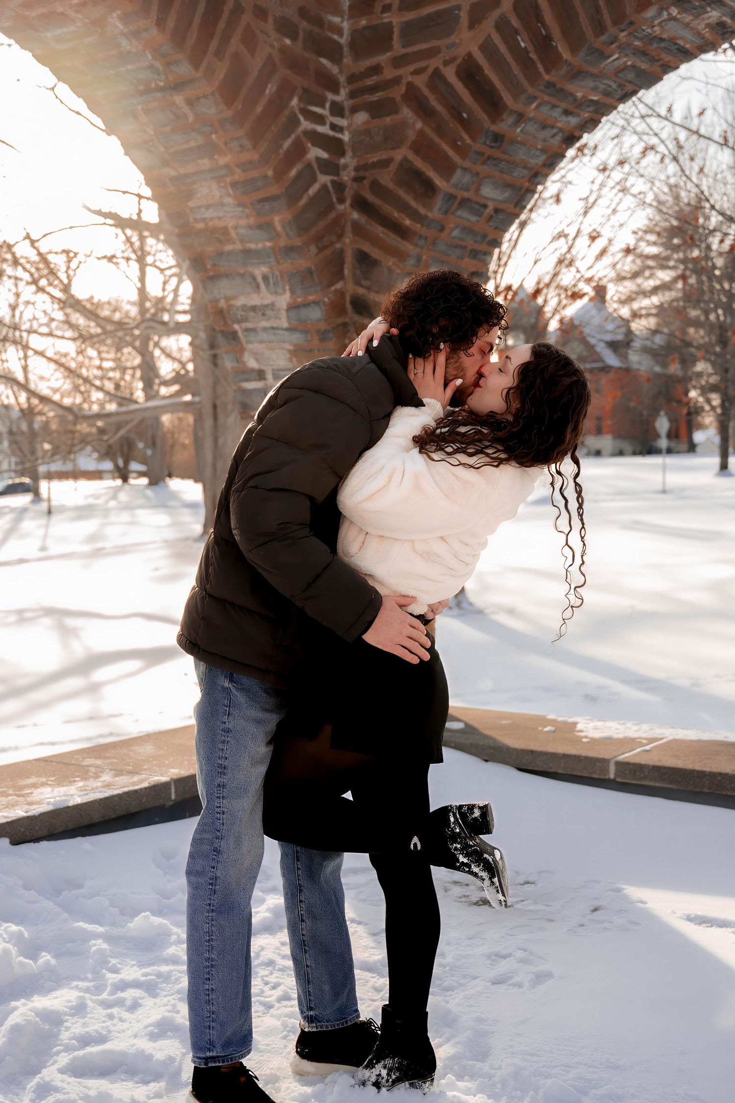 Couple kissing passionately under a brick arch surrounded by snow in winter sunlight.
