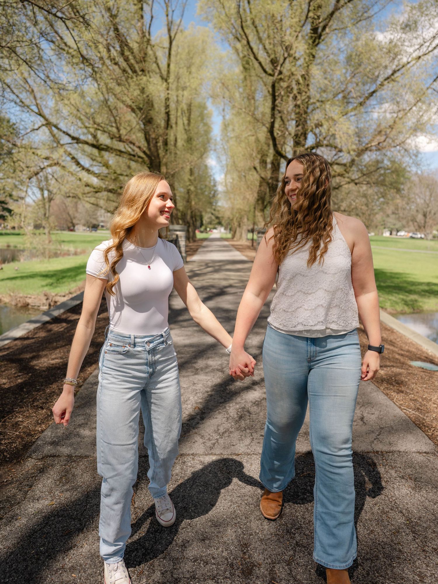 Two young women hold hands and smile while walking along a sunny tree-lined park pathway in casual jeans.