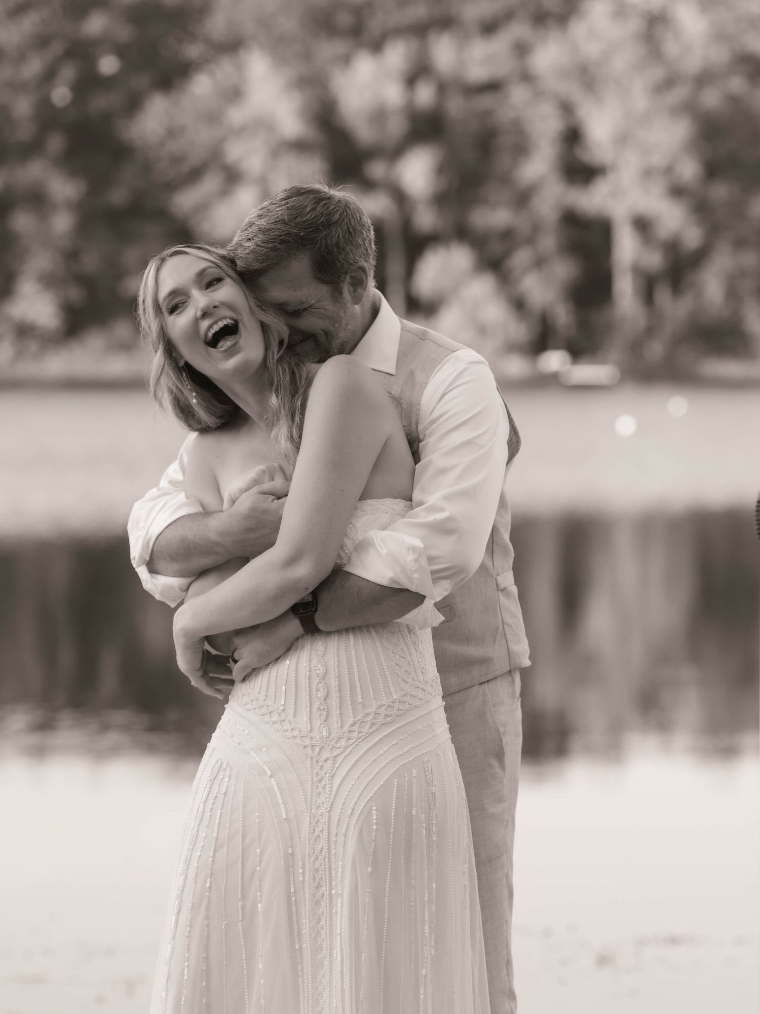 Black and white photo of a laughing bride in white lace gown embracing groom outdoors near a lake.