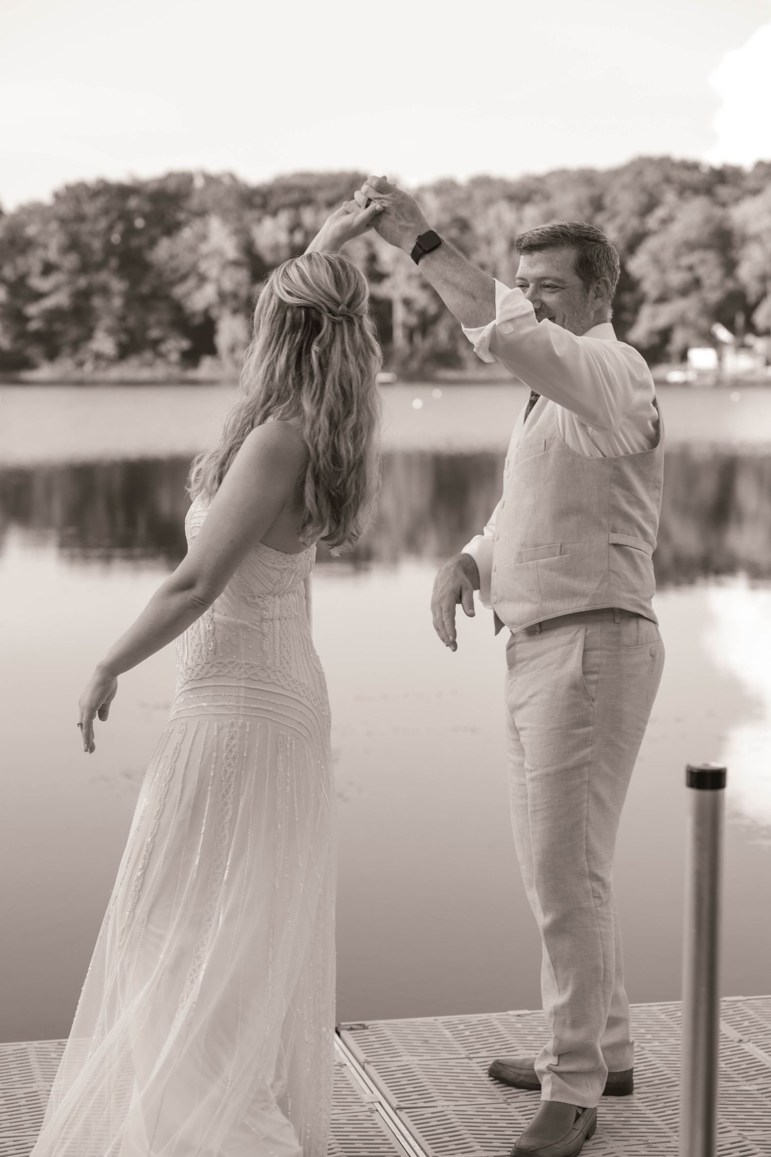 Couple dancing on a lakeside dock in black and white, bride in lace gown, groom in white linen suit.