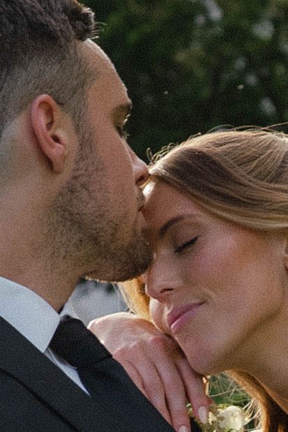 Groom tenderly kisses bride on forehead in close-up romantic wedding portrait outdoors among green foliage.