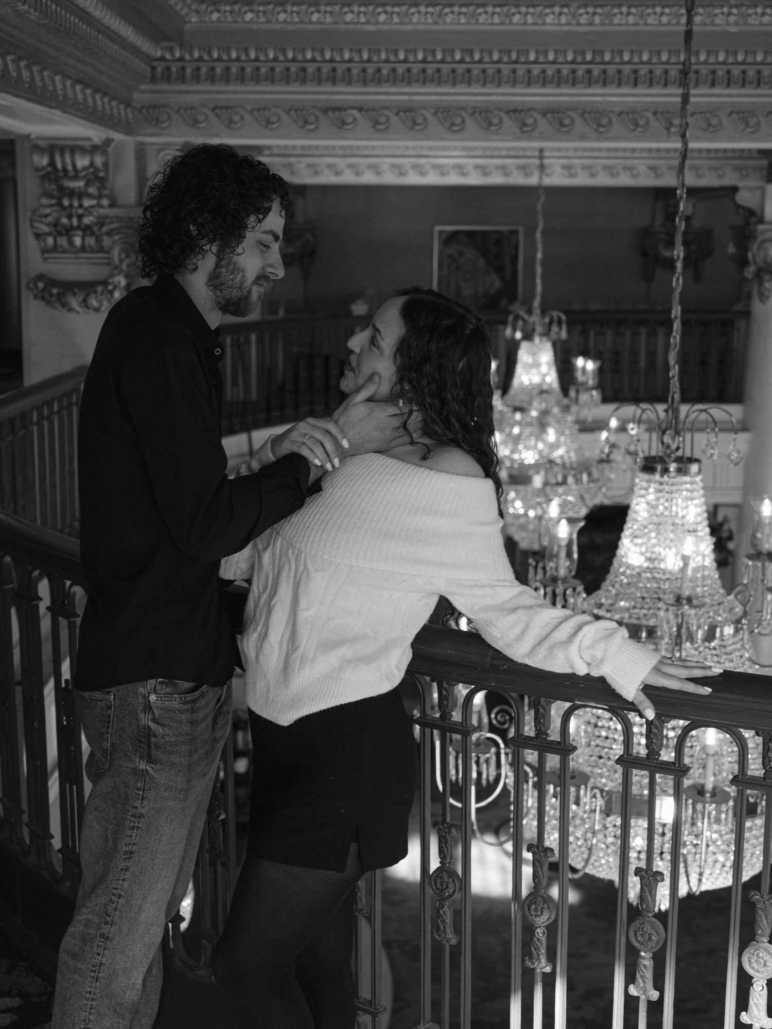 Black and white photo of couple embracing on a balcony with crystal chandeliers in a grand ballroom.
