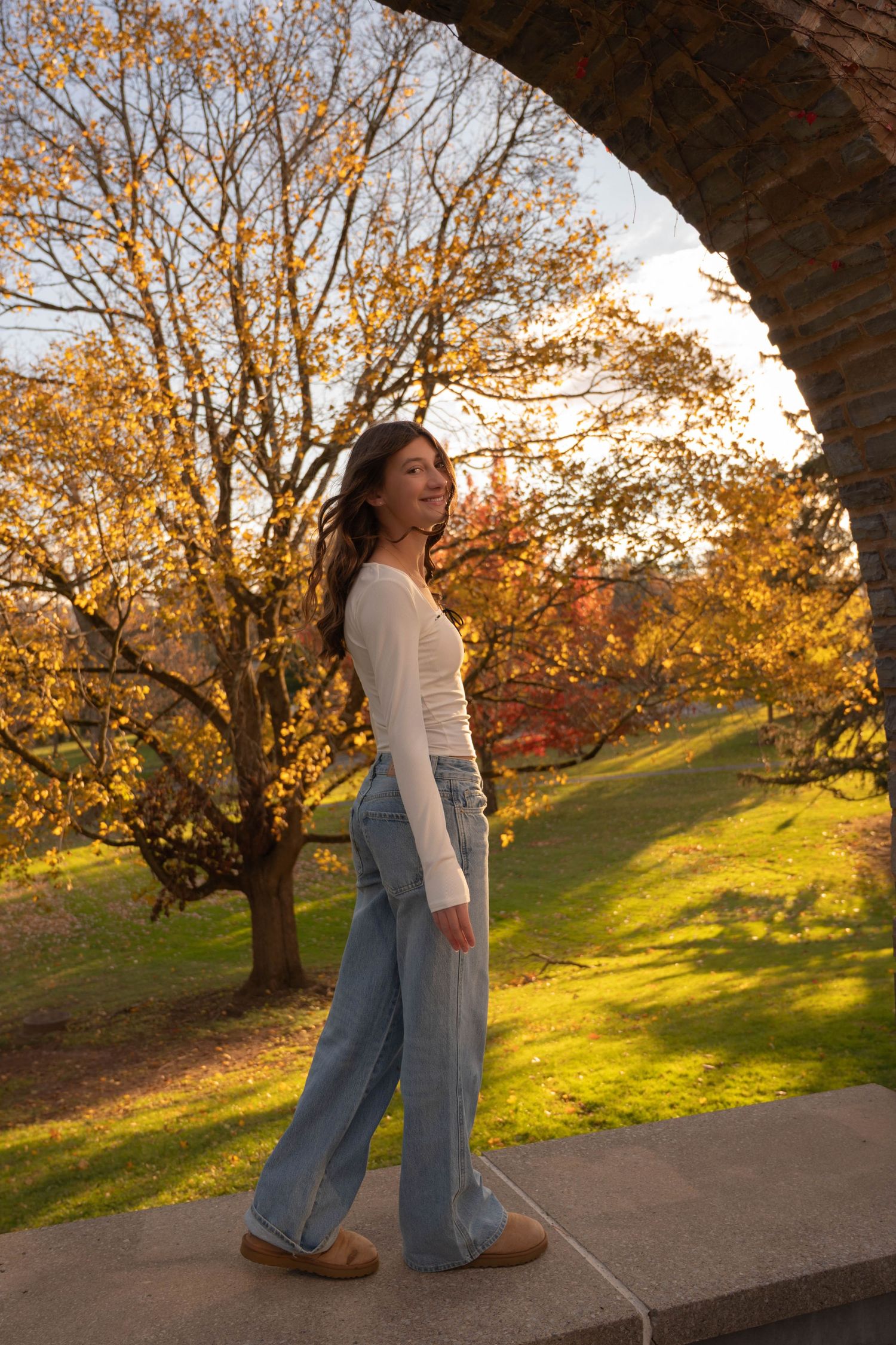 Young teen girl in white top and wide-leg jeans posing under stone arch in golden autumn park.
