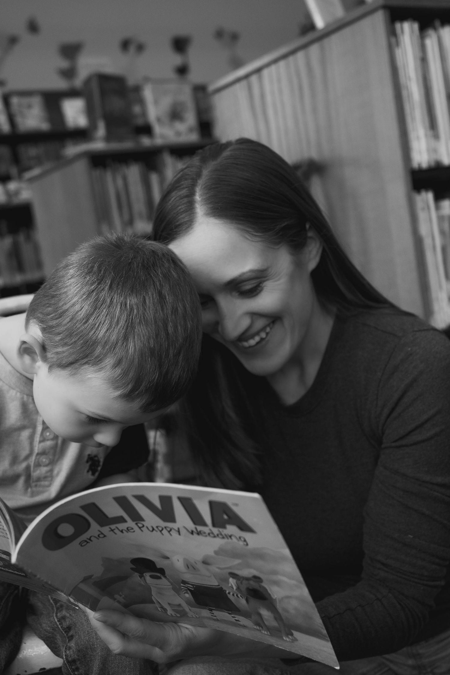 Black and white photo of a smiling mother and son reading the book Olivia together in a library.