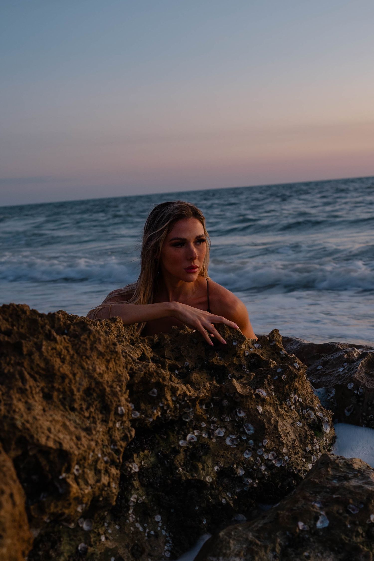 Blonde woman peers over moss-covered rocks on beach during dusky twilight ocean scene.