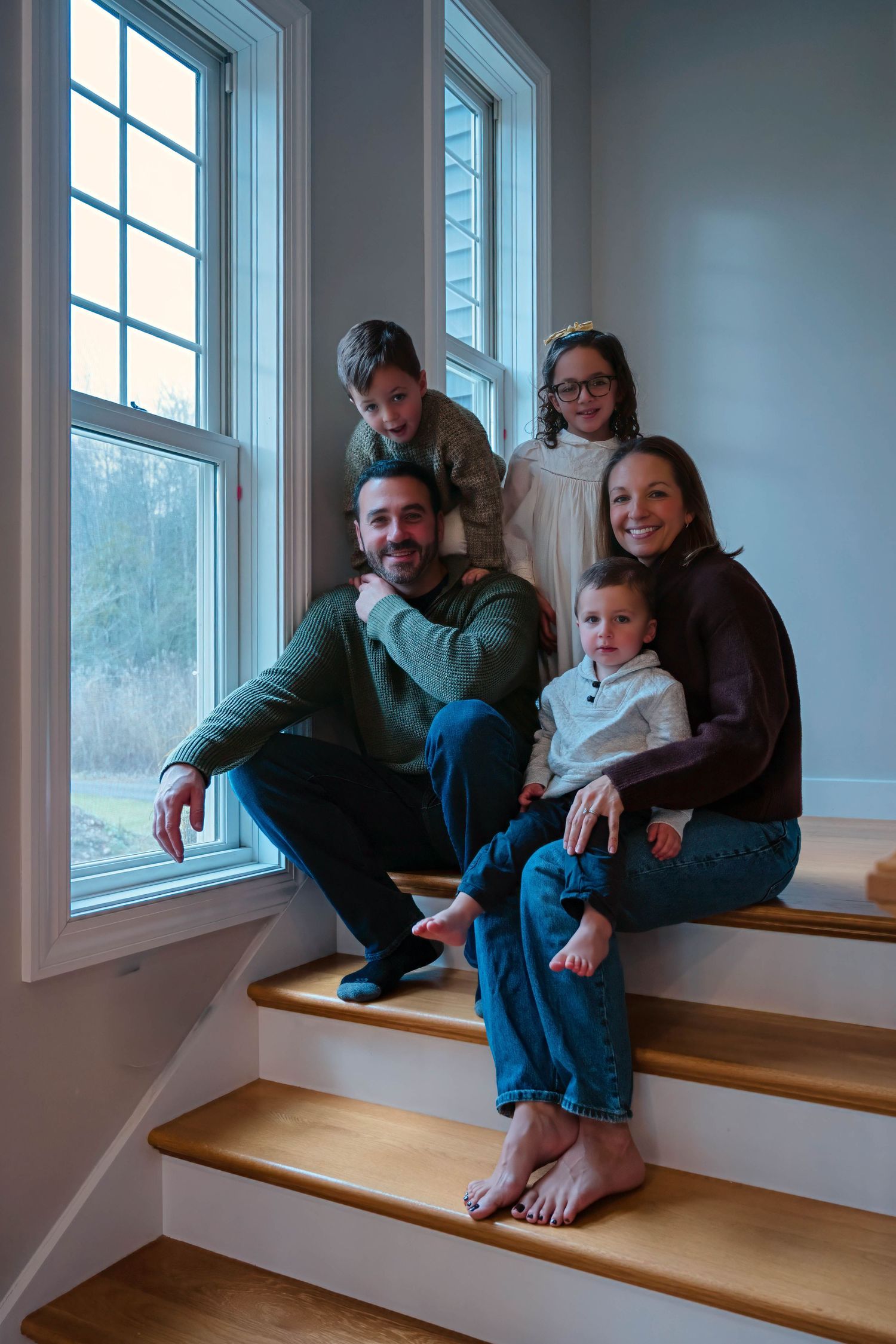 Smiling family of five poses together on wooden stairs near large windows in a bright home.