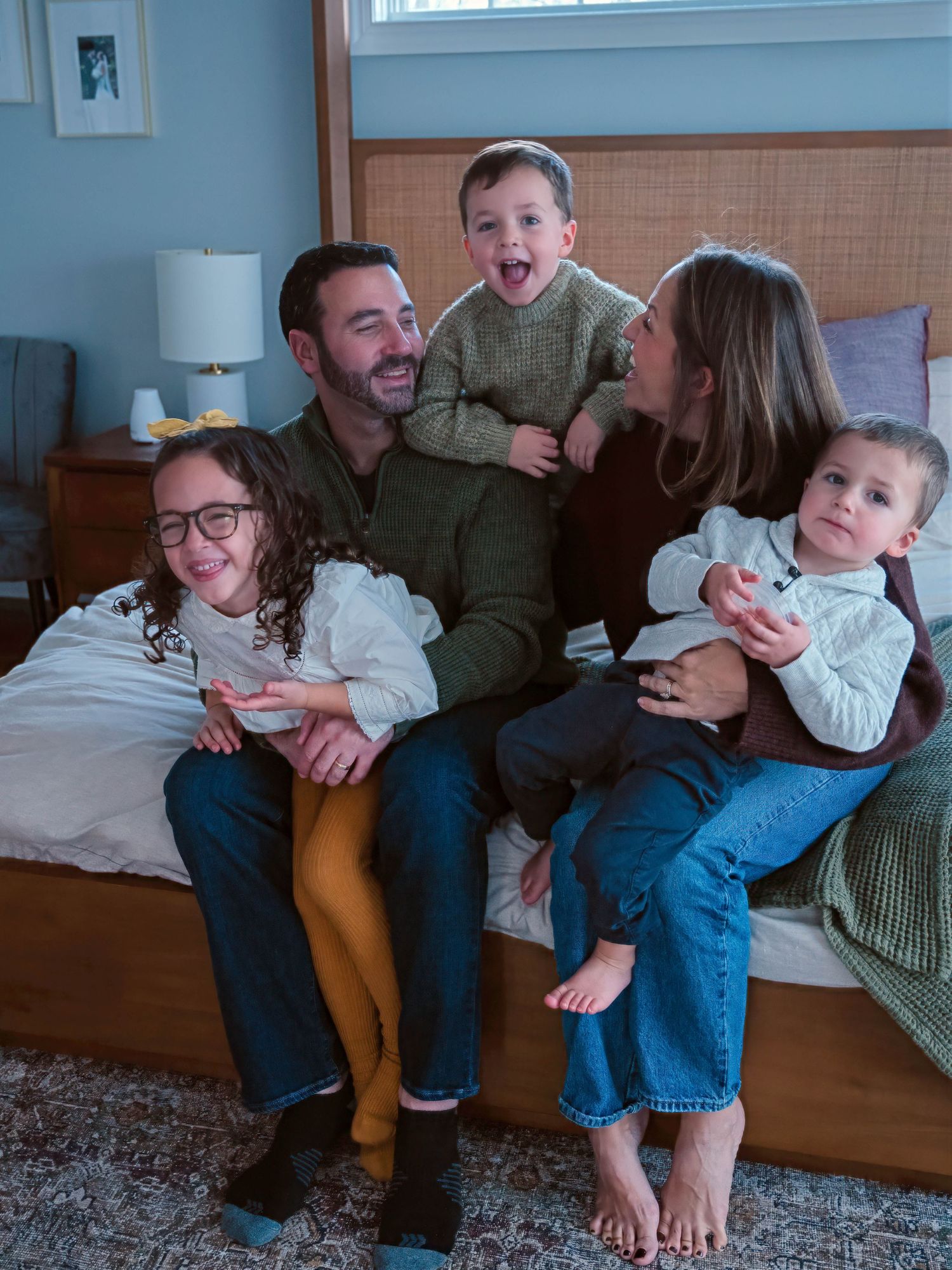 A joyful family of five sits together on a bed, laughing and sharing a fun moment.