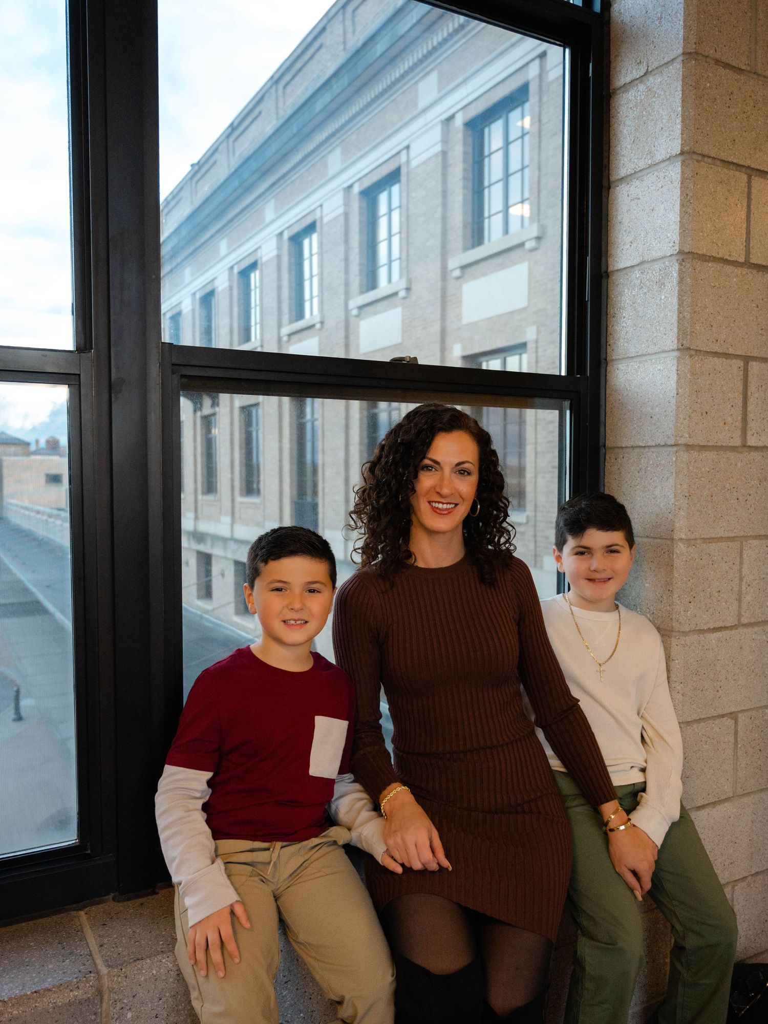Pregnant woman in brown dress sits with two boys by large windows in elegant building interior.