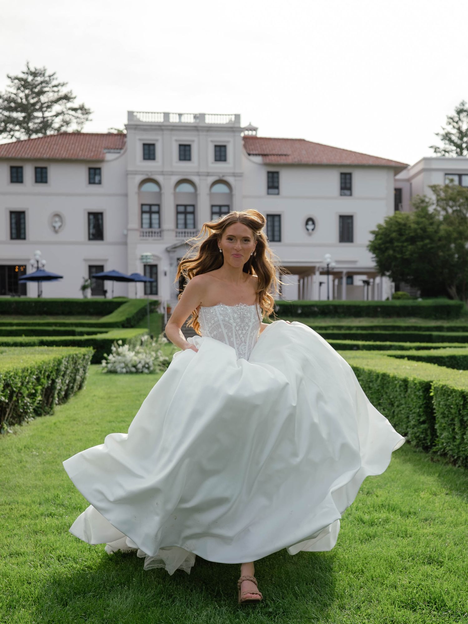 Bride in strapless corset ball gown twirling on green lawn in front of grand white wedding venue.