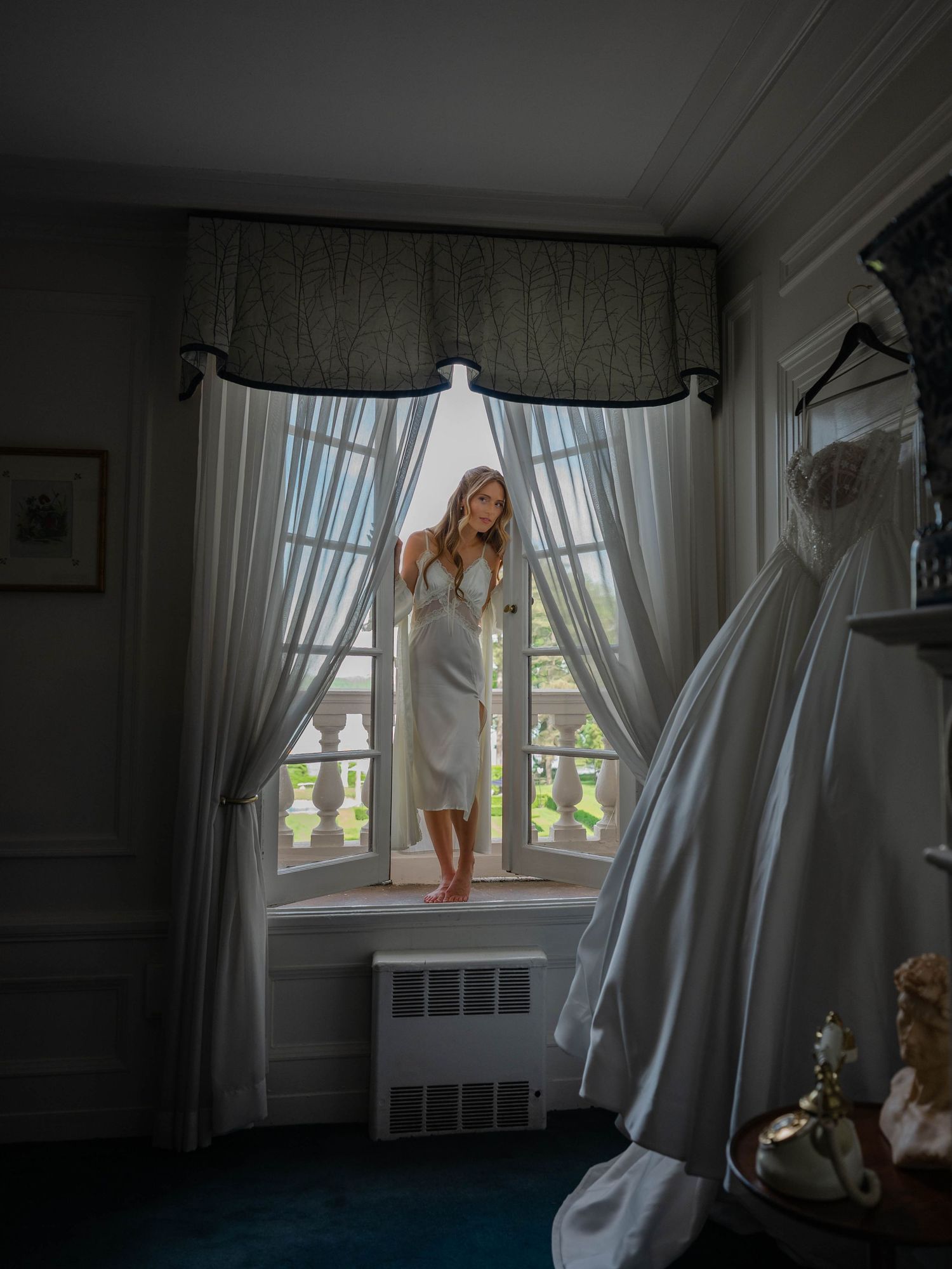 Bride in white slip dress stands barefoot in bay window beside hanging wedding gown in elegant room.