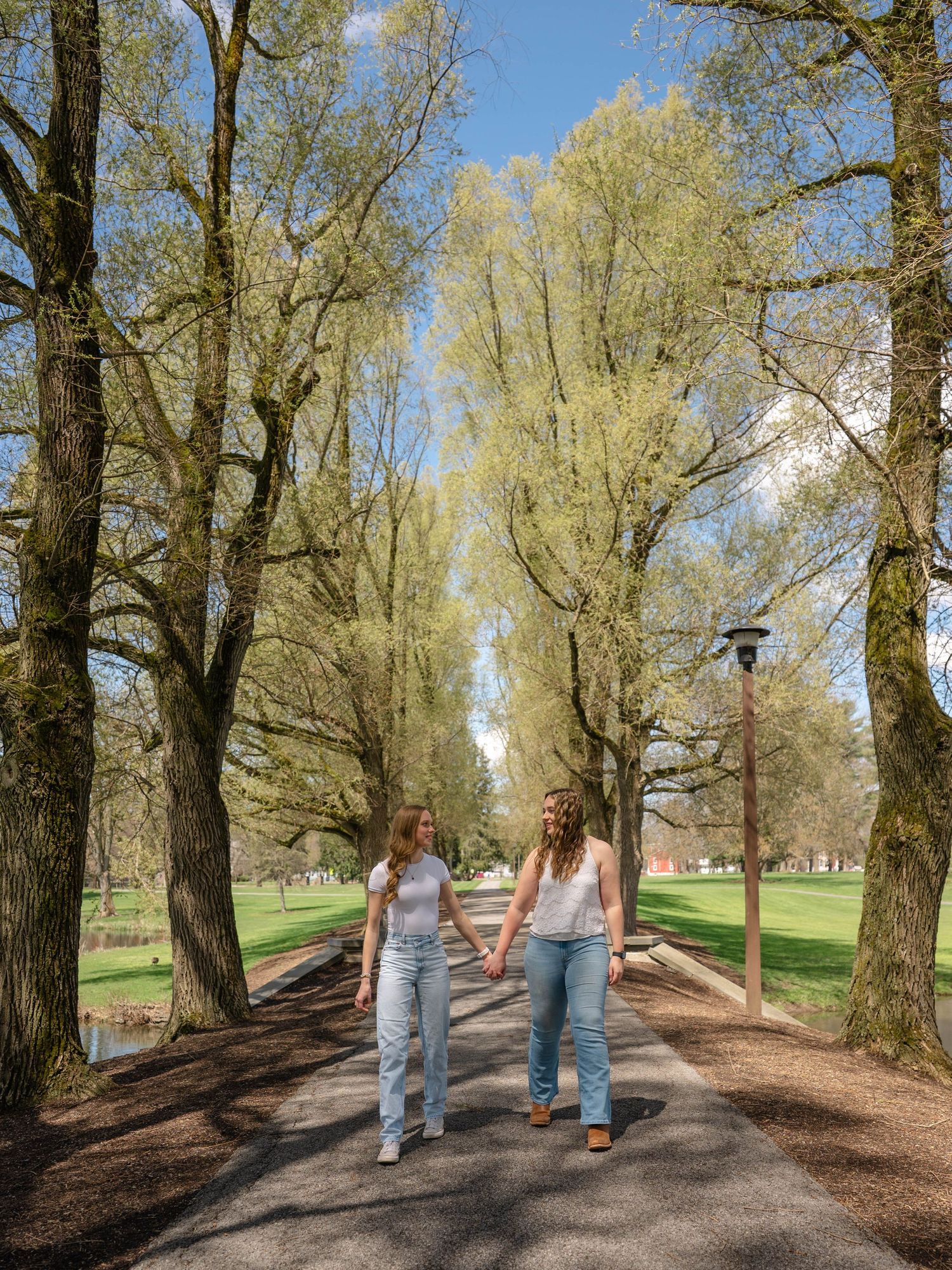 Two young women hold hands while walking on a scenic tree-lined path under a bright blue sky.