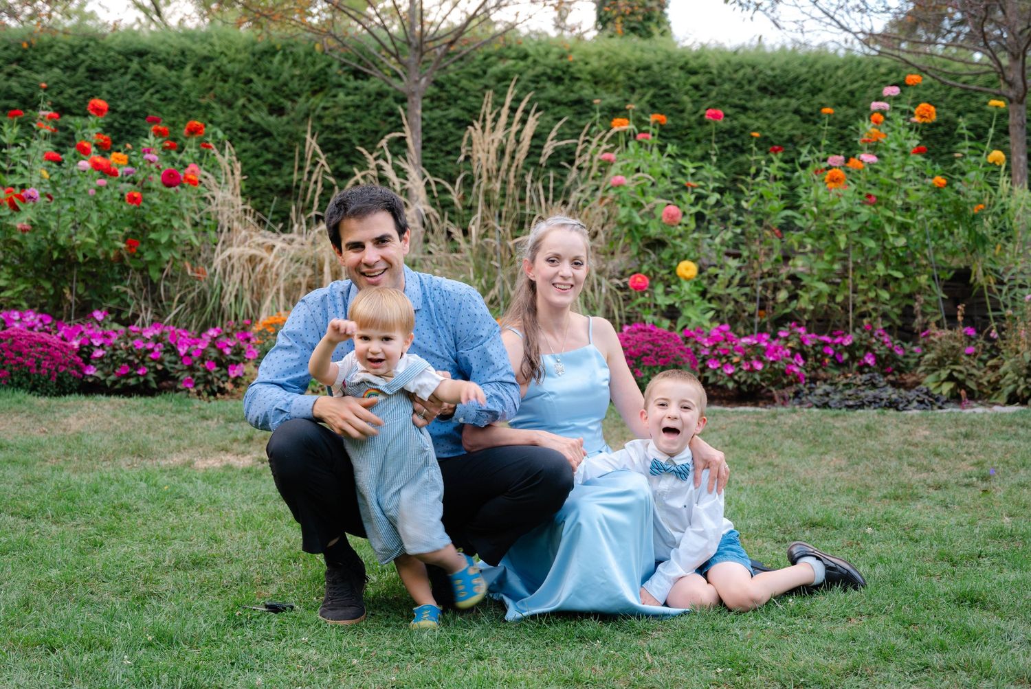 Happy family of four poses in a colorful flower garden, parents with two young children on green grass.