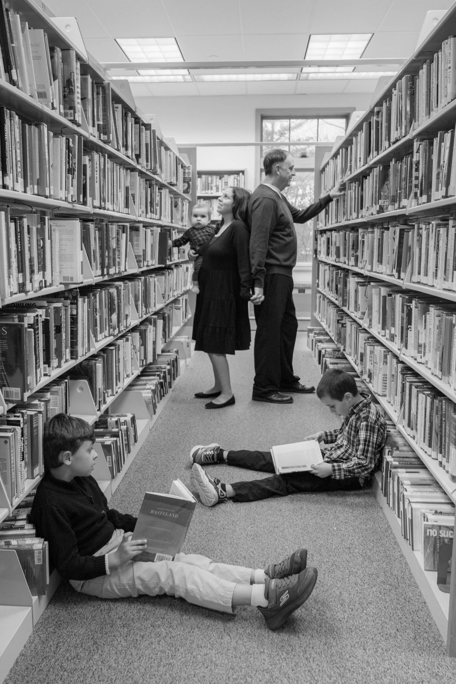 Black and white photo of family browsing books in library aisle, two boys reading on floor.