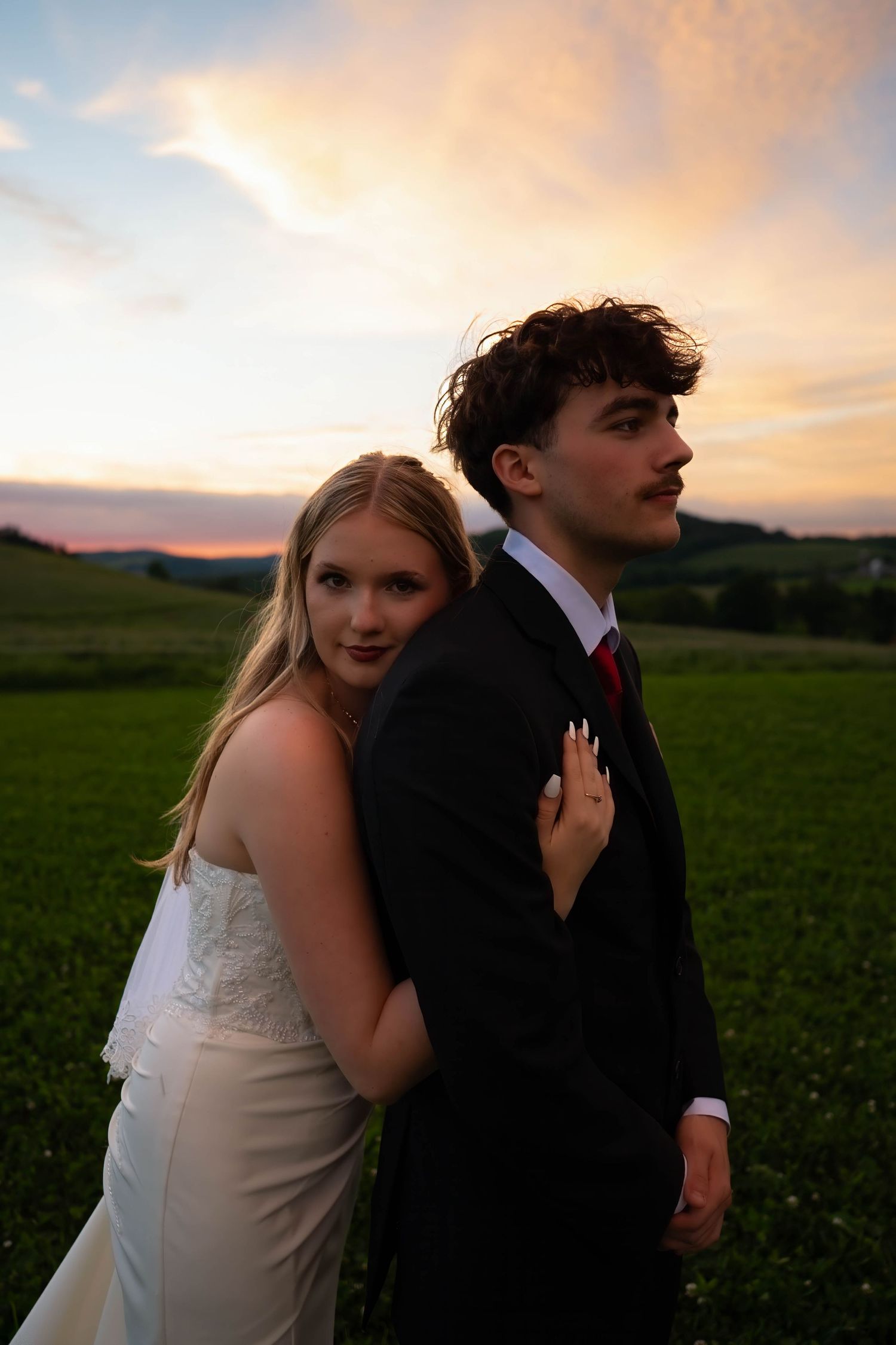 A young couple in formal attire poses at sunset in a green field, woman in white dress embracing man in black suit.