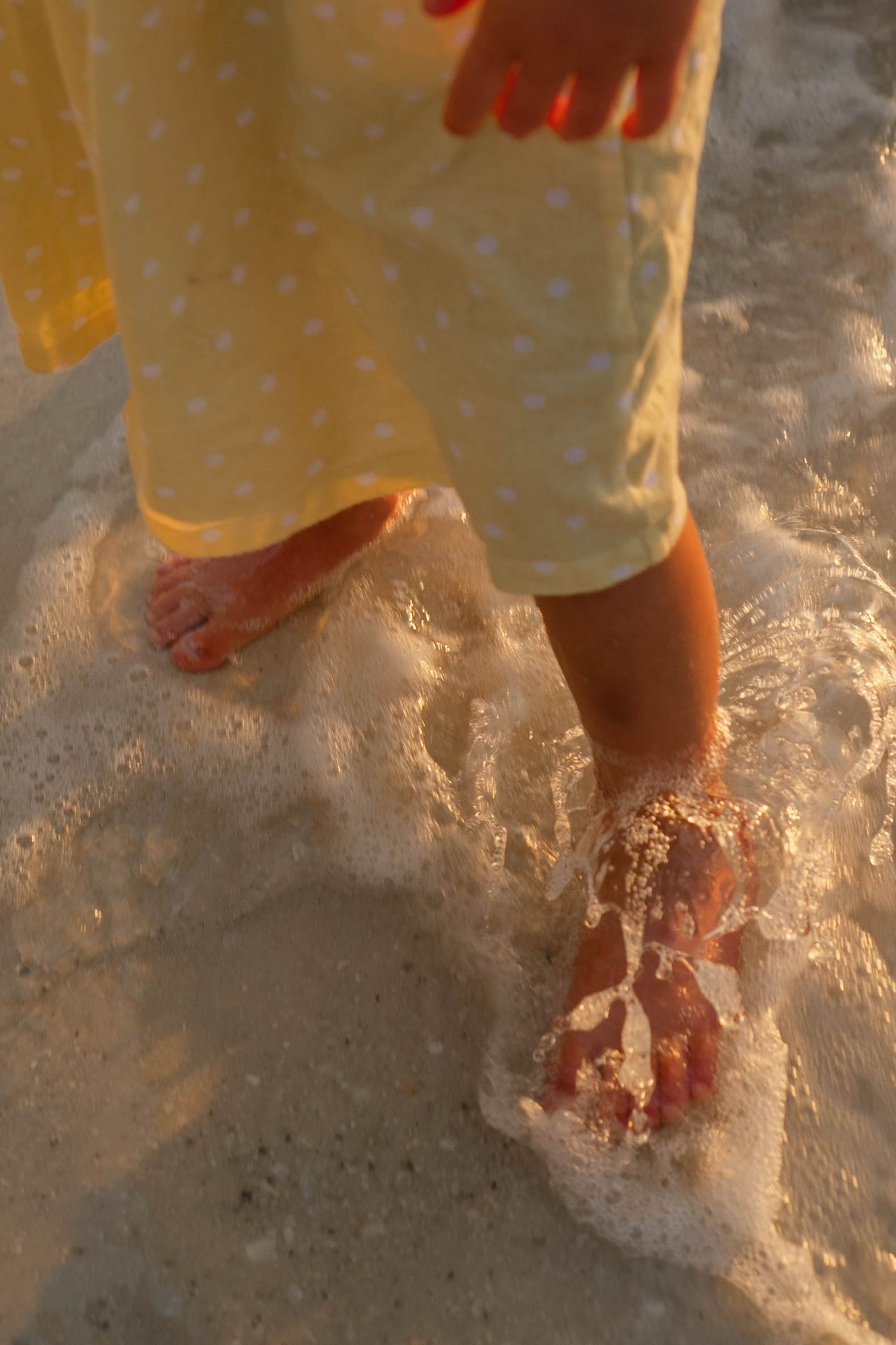 Child's bare feet in ocean waves on sandy beach, wearing yellow polka dot dress at sunset.