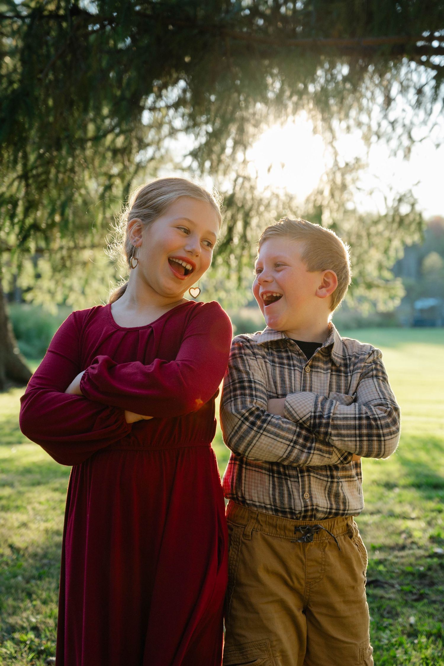 Two laughing children with arms crossed pose outdoors in golden sunlight, girl in red dress and boy in plaid shirt.