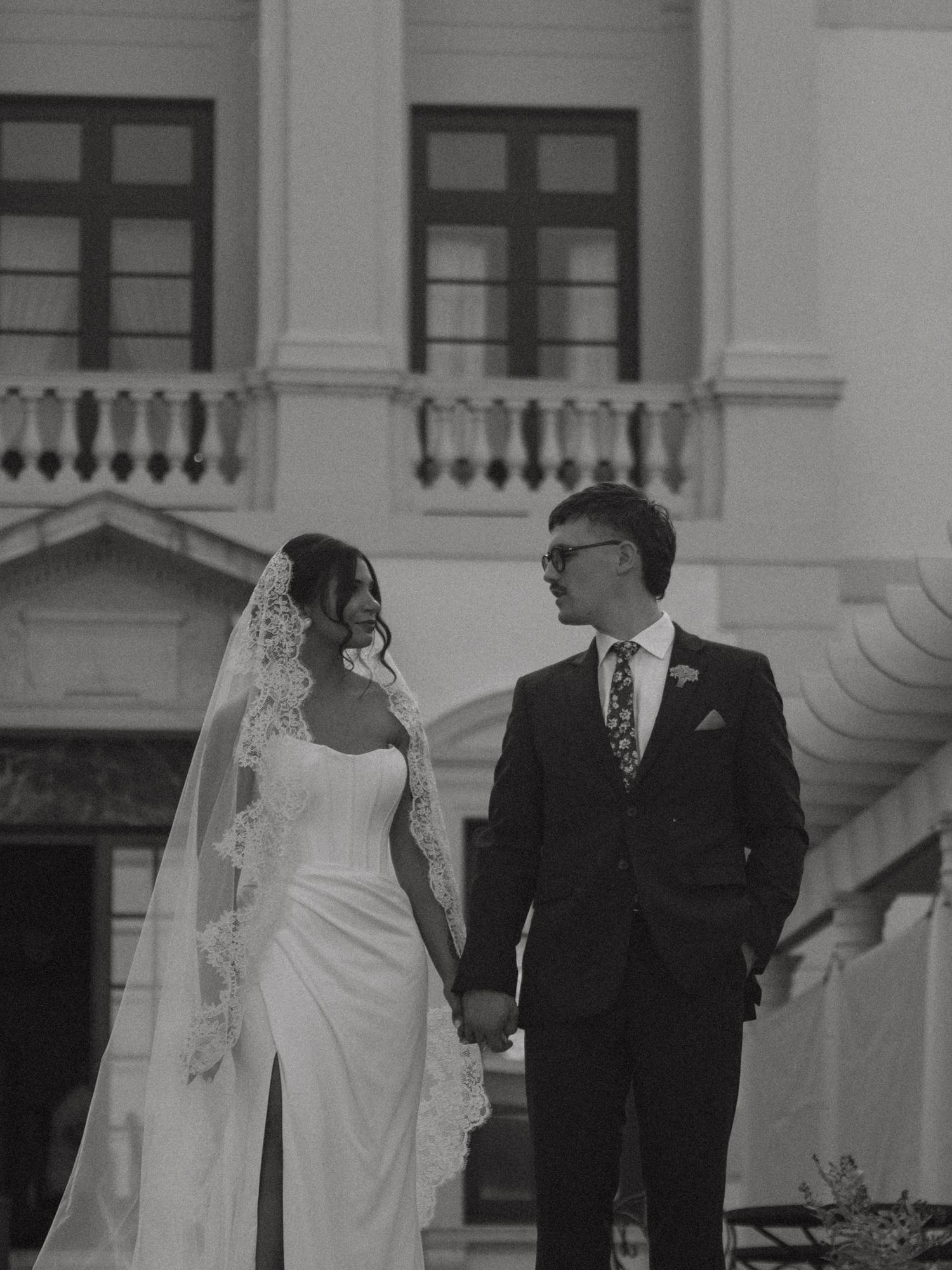 Black and white wedding portrait of bride in lace veil and groom holding hands outside classic building.