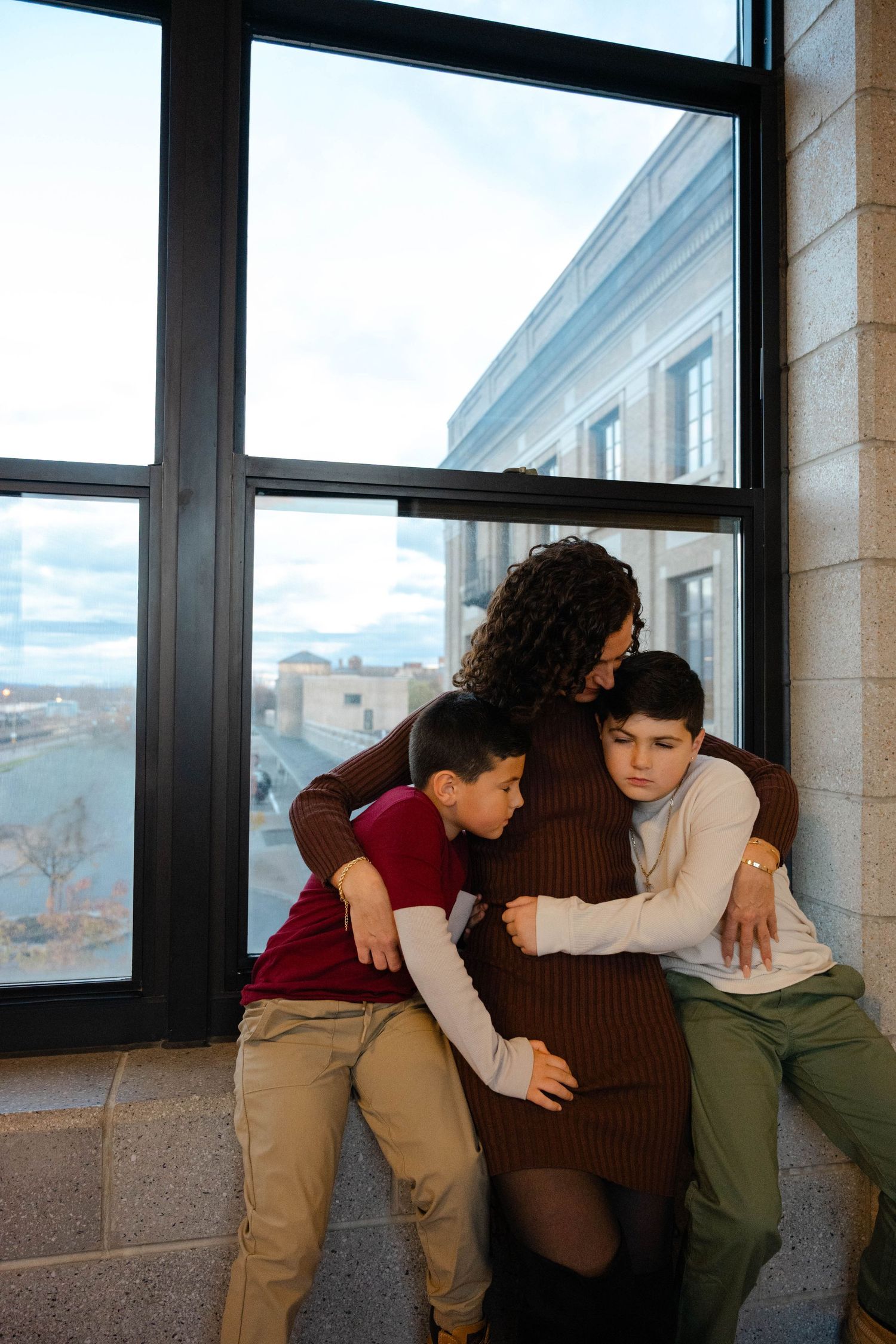 Two young boys lovingly hug their pregnant mother while seated at a window ledge indoors.