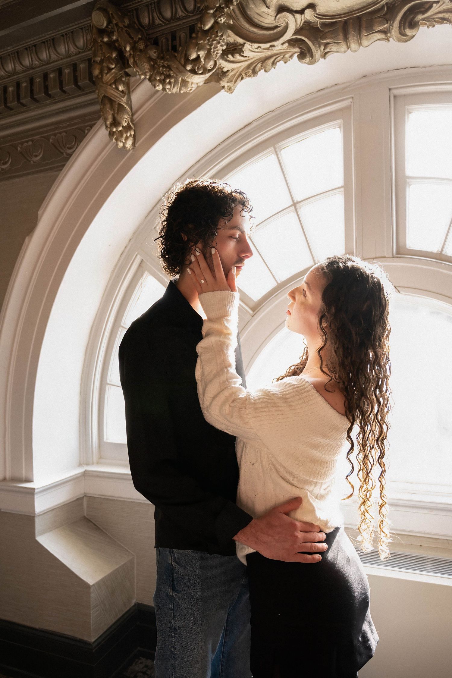 A couple embraces tenderly beneath an ornate arched window, gazing lovingly at each other.