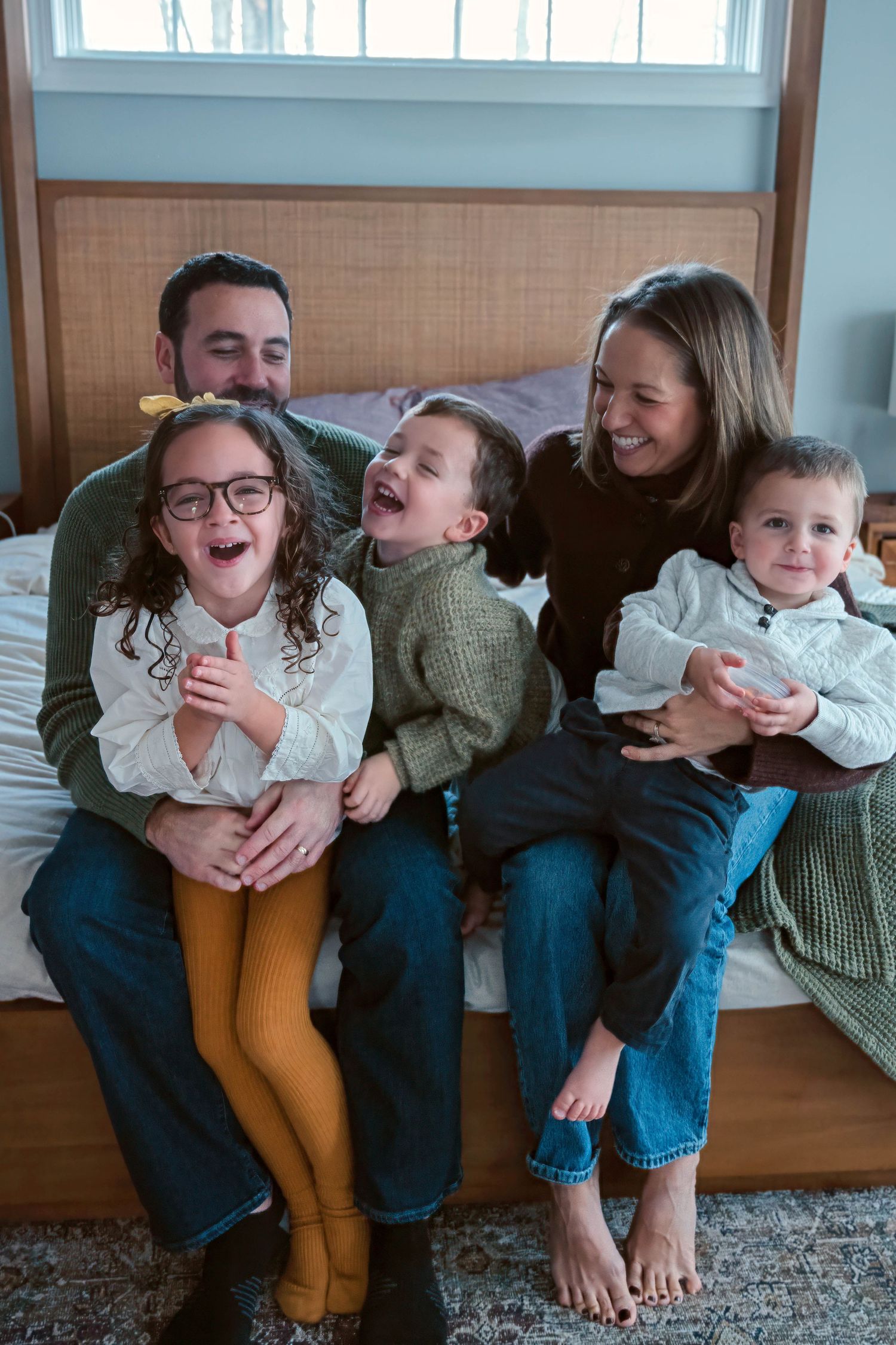 Happy family of five laughing together on a bed, parents with three young children in cozy home setting.
