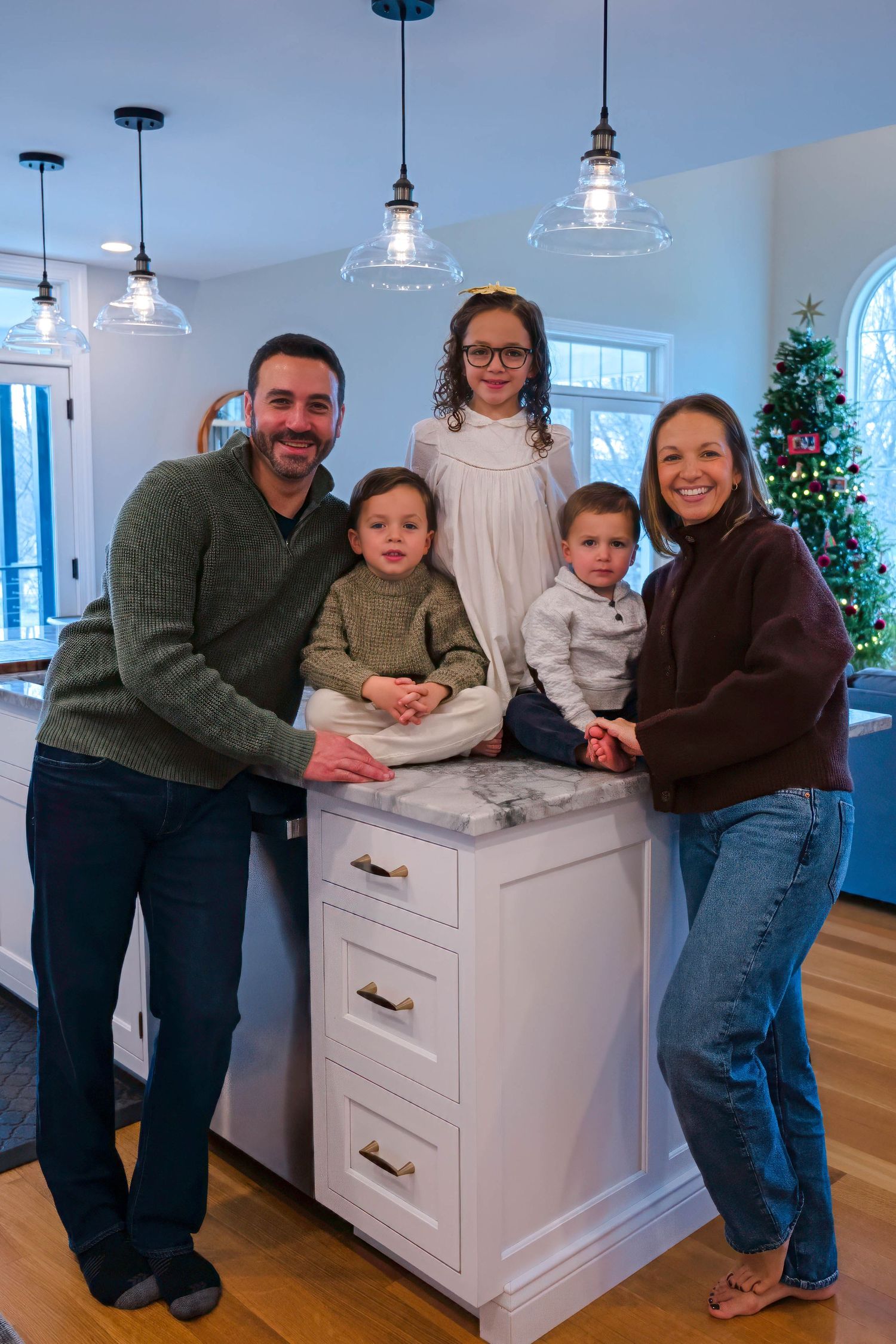 Family of five smiles together around white kitchen island with Christmas tree in background.