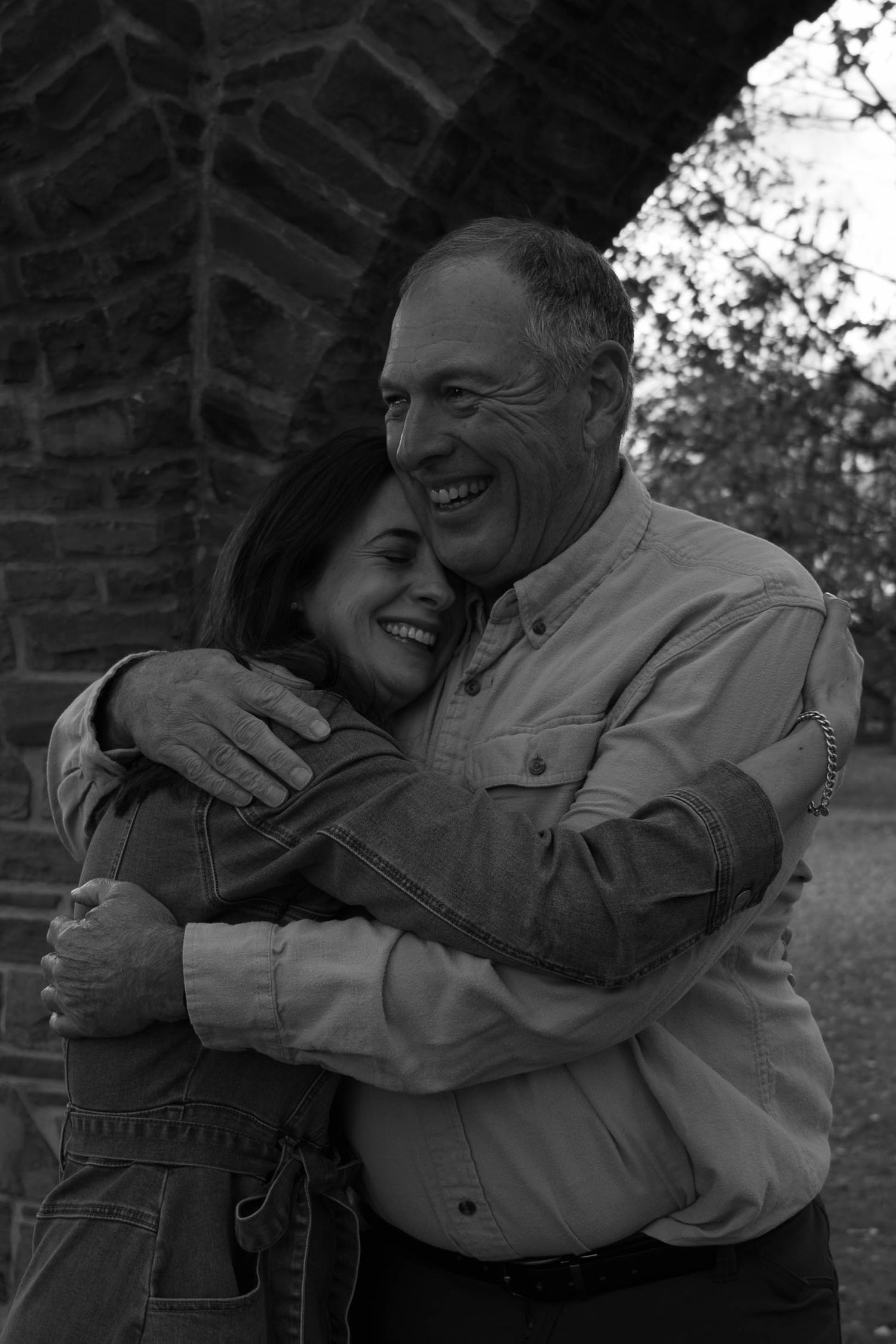 Black and white photo of a smiling older man and woman hugging warmly near a stone arch outdoors.