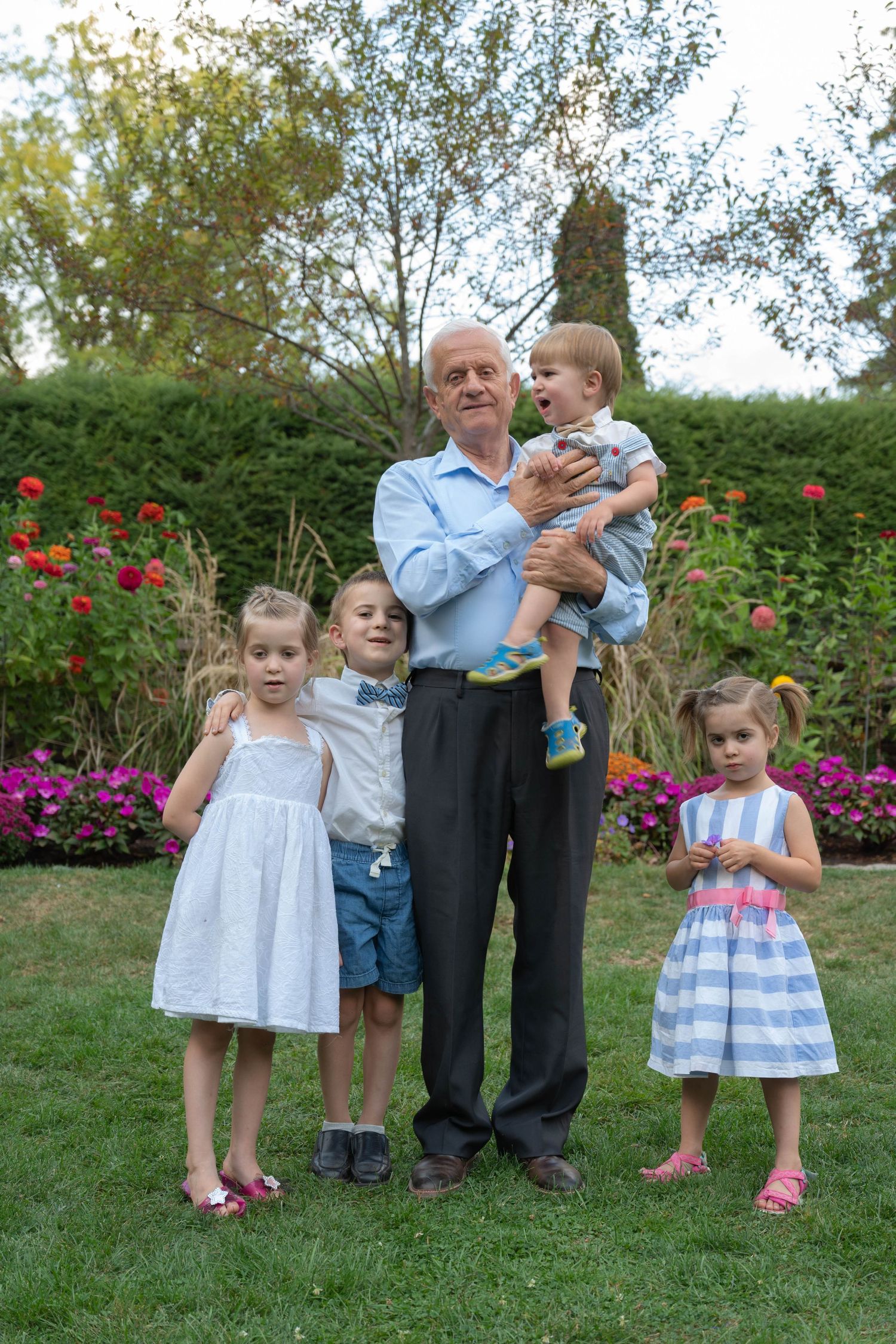 Elderly grandfather holding a toddler while three young children stand beside him in a beautiful garden setting.