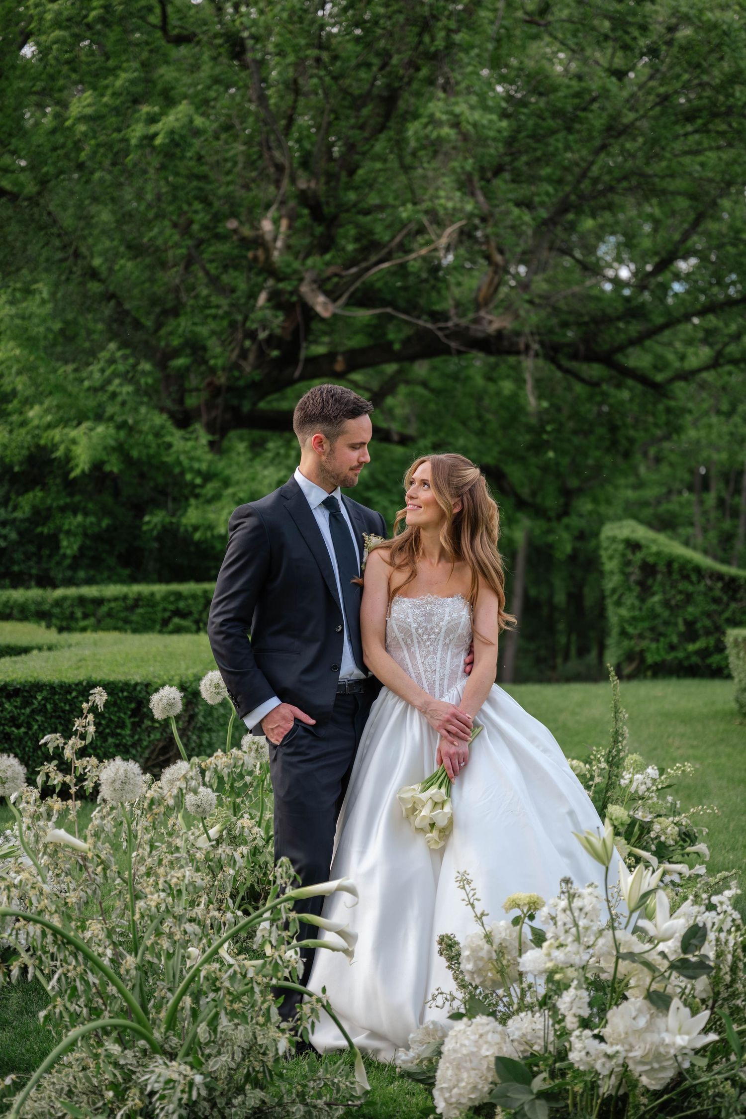 Bride and groom stand closely together among white garden flowers, gazing at each other in lush green setting.