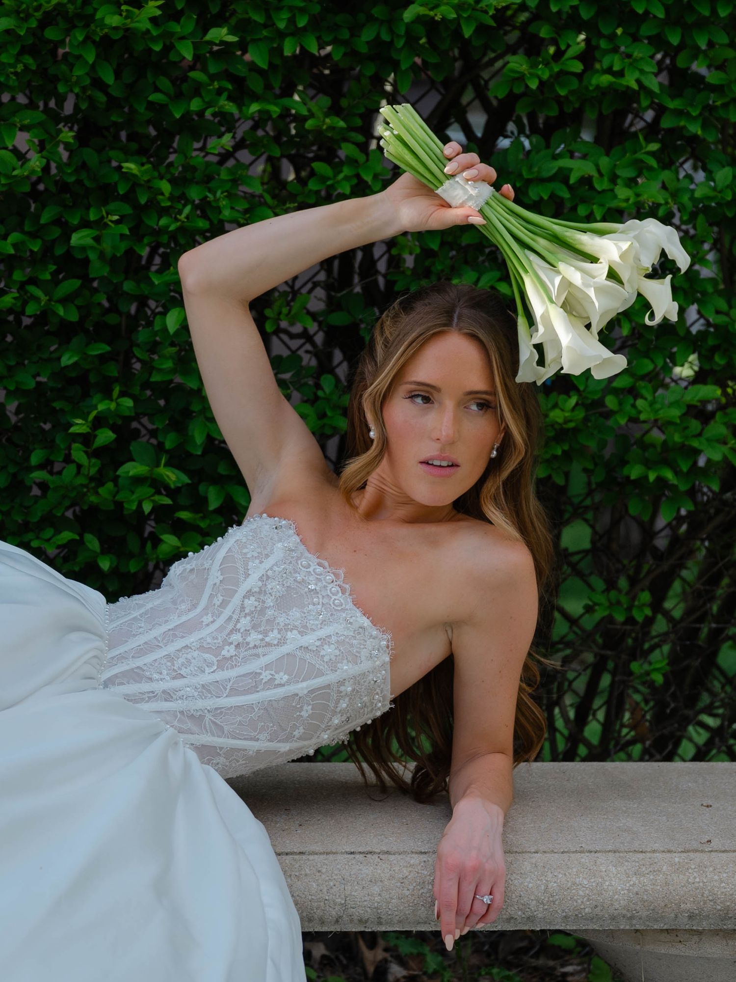 Bride in strapless lace gown poses dramatically on a stone bench holding white calla lilies overhead.