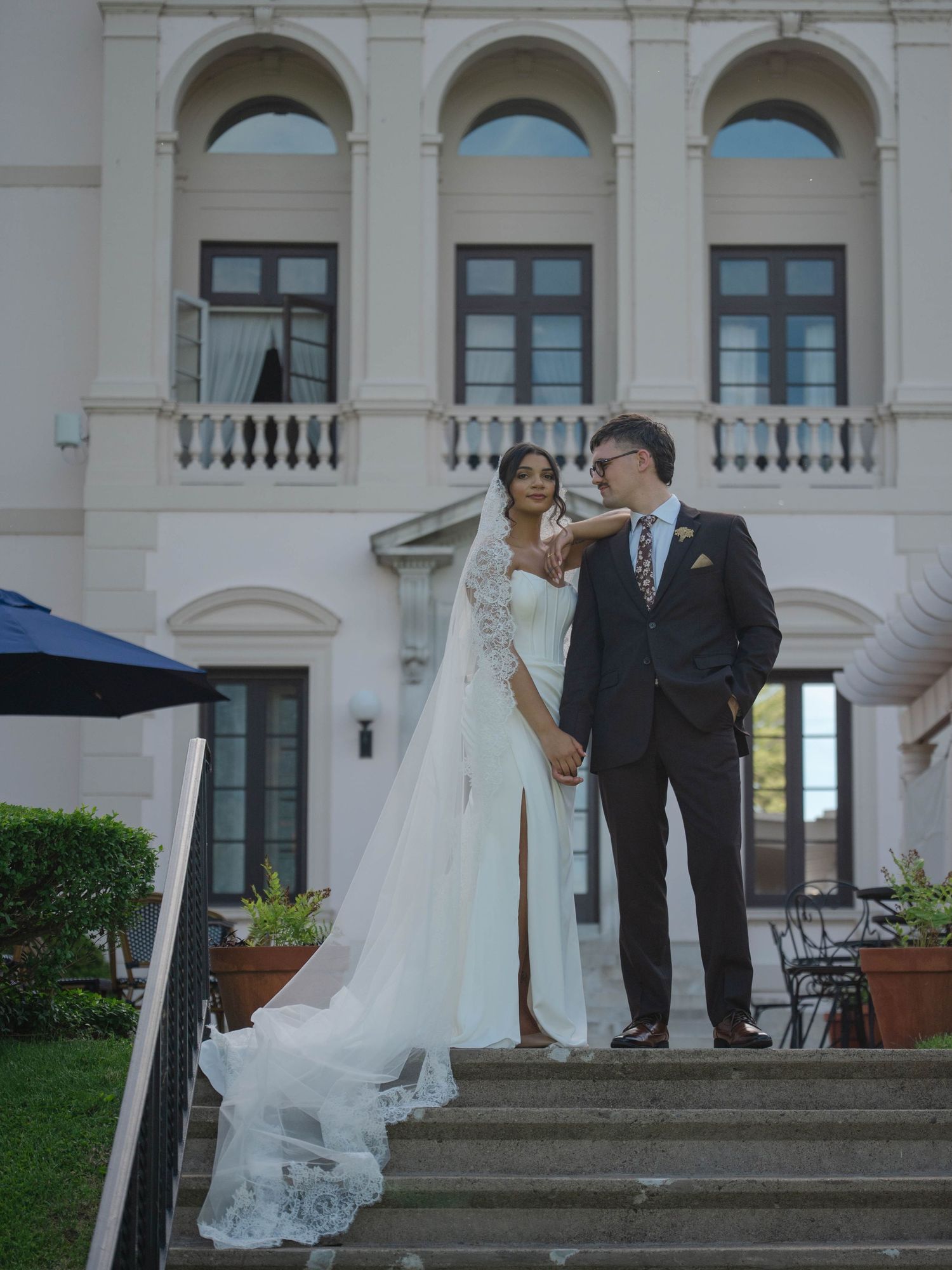 Bride and groom standing on steps of a grand white European-style villa, bride wearing a stunning lace-trimmed cathedral veil.