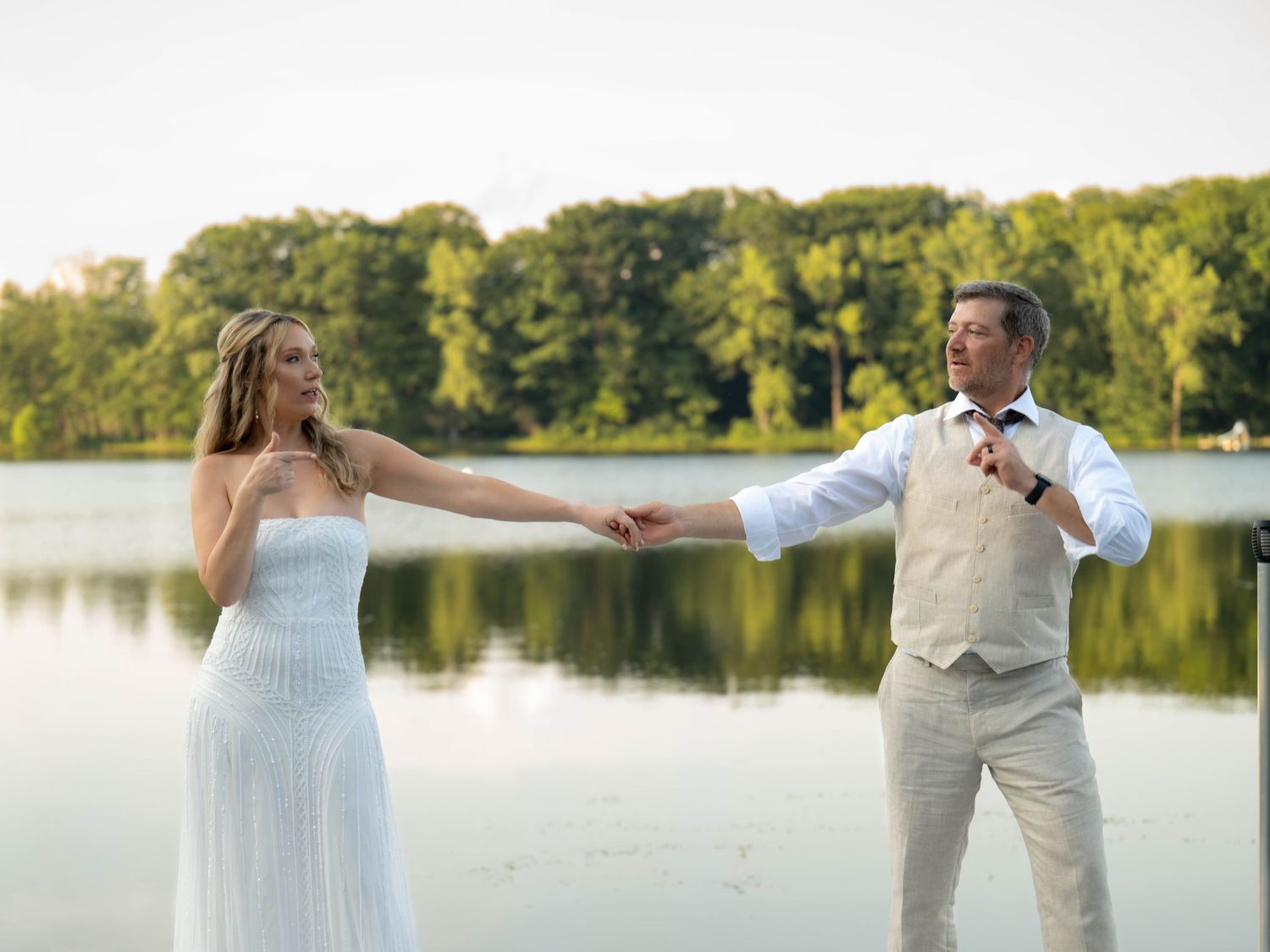 Bride in white dress and groom in vest hold hands dancing by a serene lake surrounded by lush green trees.