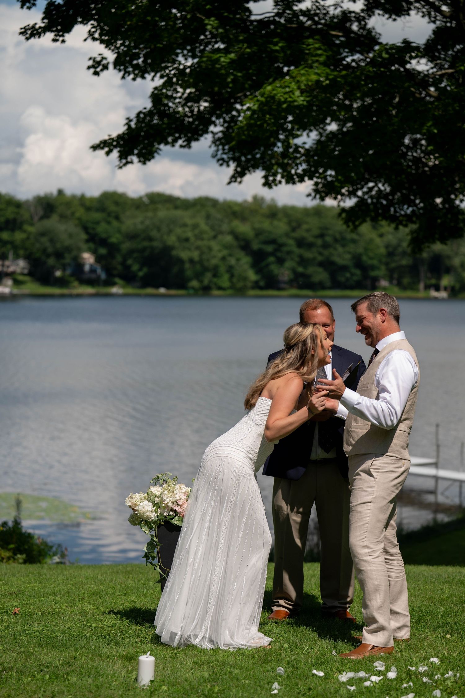 Outdoor lakeside wedding ceremony with bride in white gown and groom in tan suit, officiated under a tree.