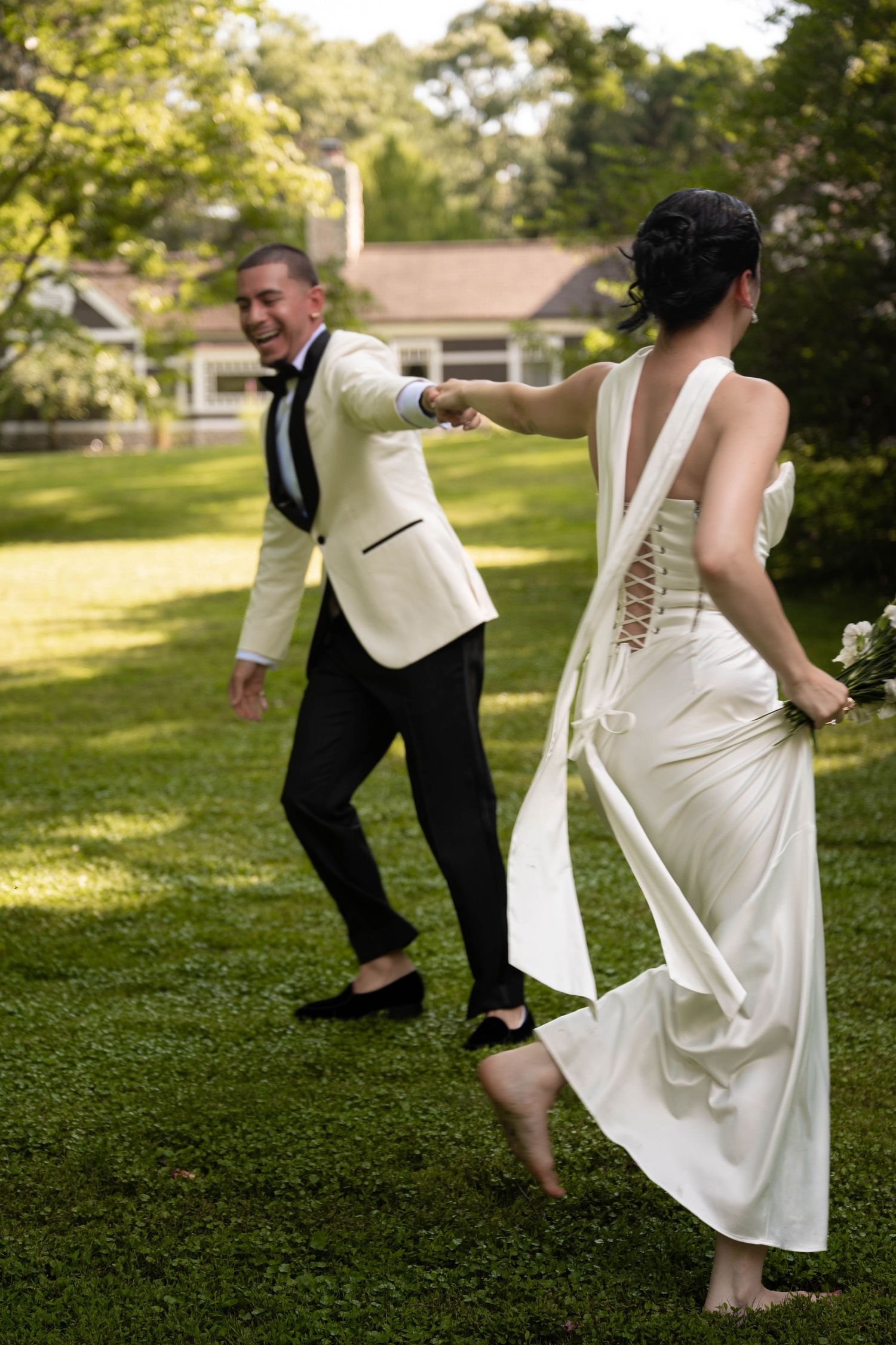 Bride and groom laughing and holding hands while running barefoot across green lawn in wedding attire.