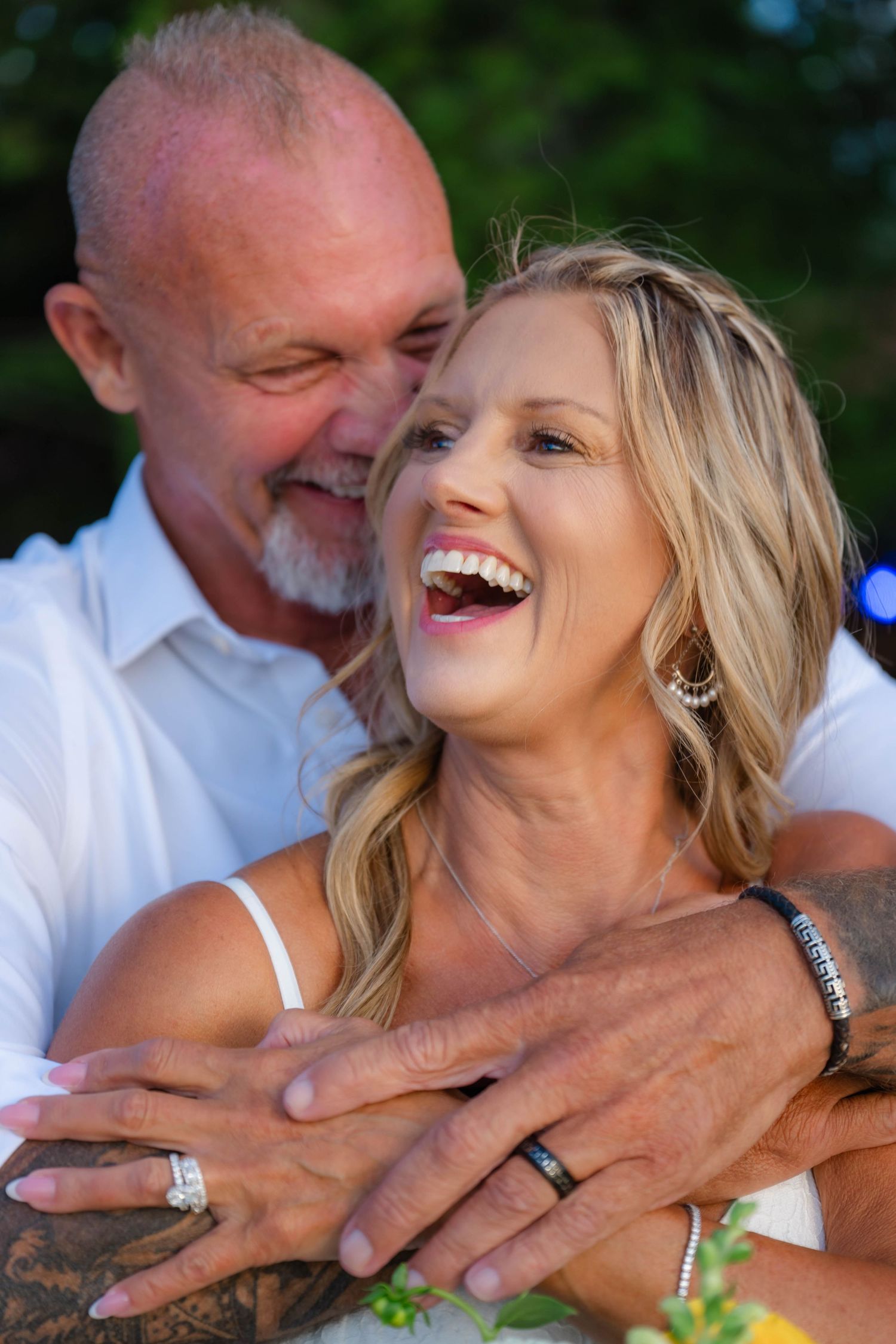 Happy couple embracing outdoors, showing wedding rings, laughing joyfully in a romantic portrait photo.
