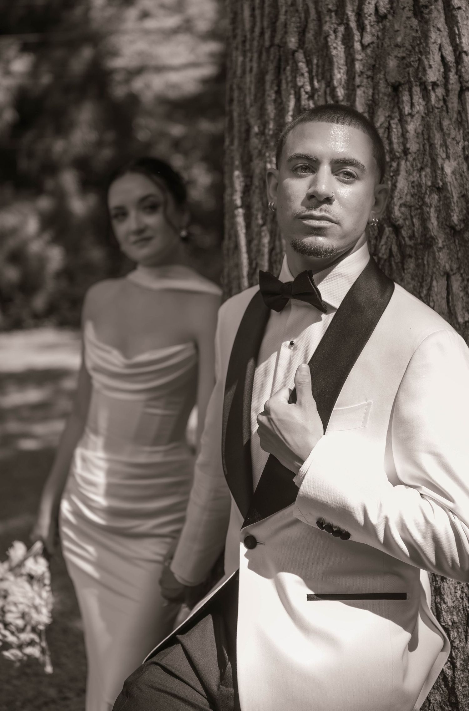 Black and white photo of a groom in white tuxedo leaning on tree with bride in strapless gown in background.
