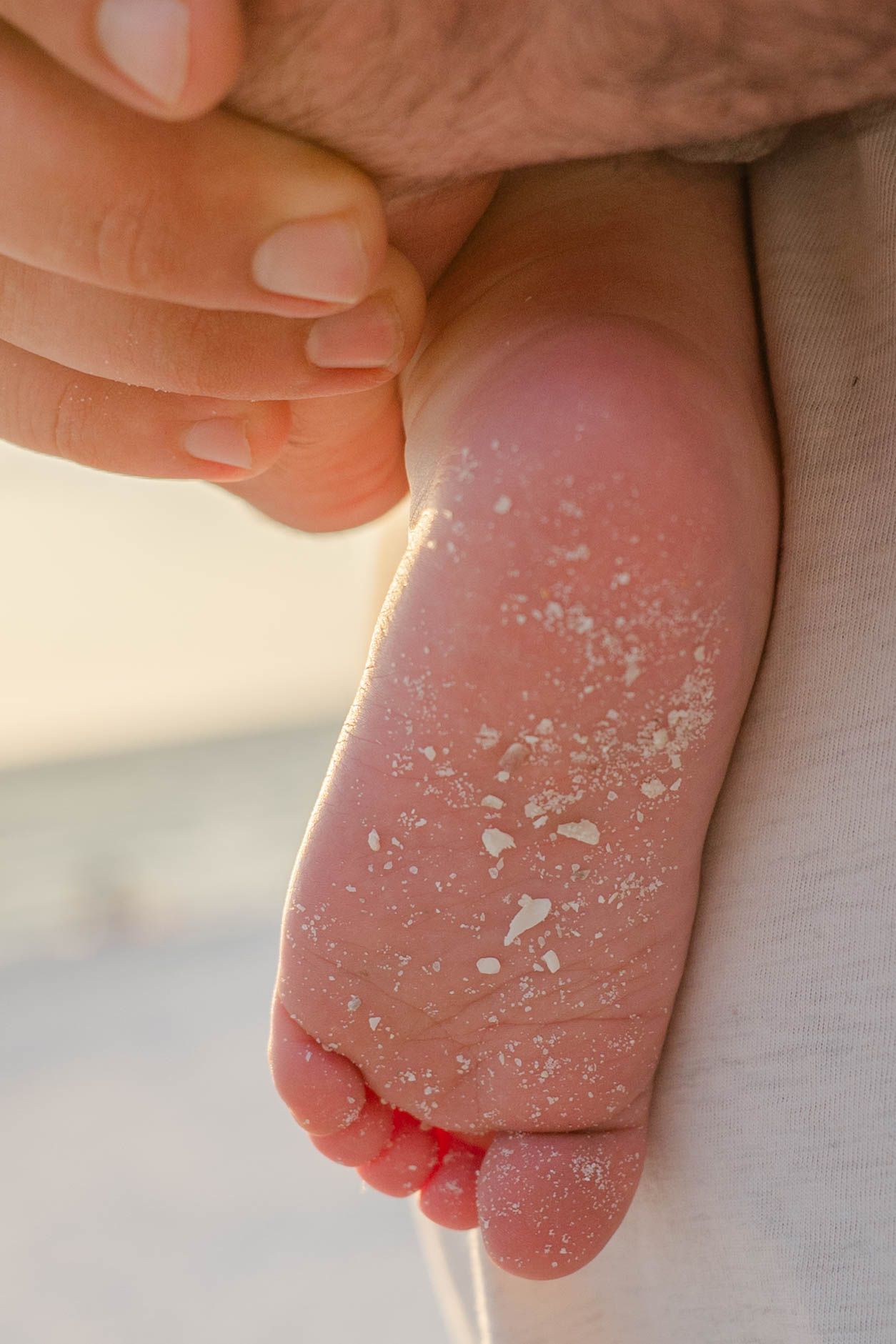 Close-up of a sandy baby foot being held by an adult hand at the beach.