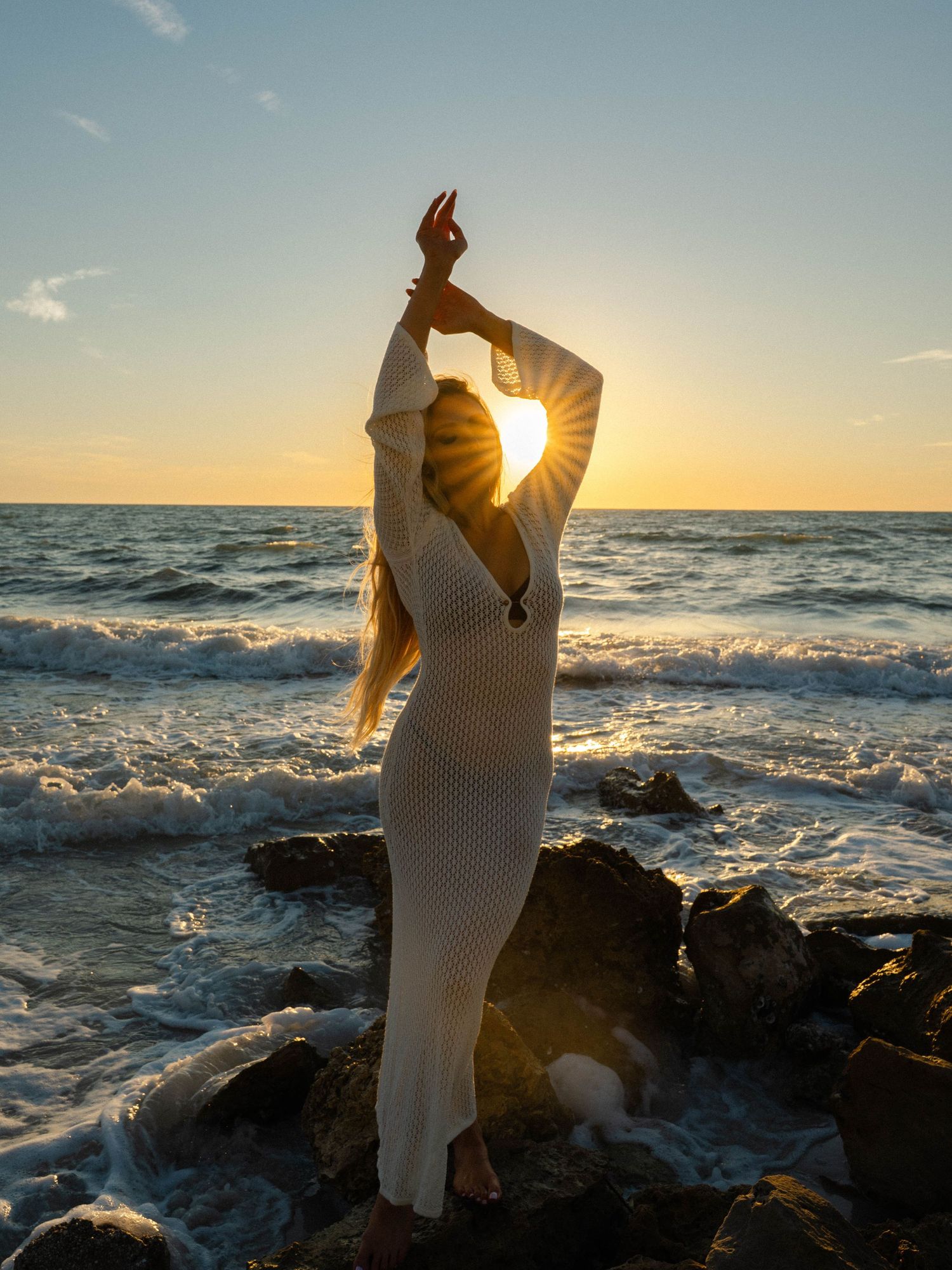 Woman in white crochet dress poses on rocky beach at golden sunset with arms raised, sun rays bursting through her hands.