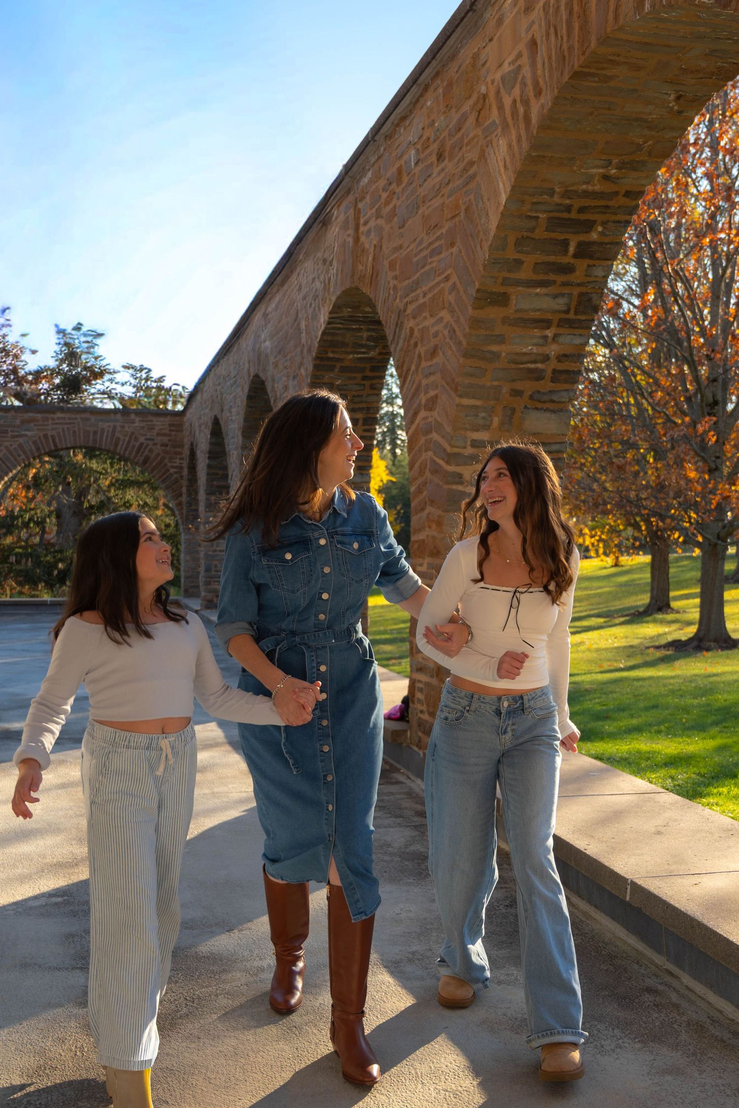Three young women walk and laugh together under a stone arch bridge surrounded by autumn trees on a sunny day.