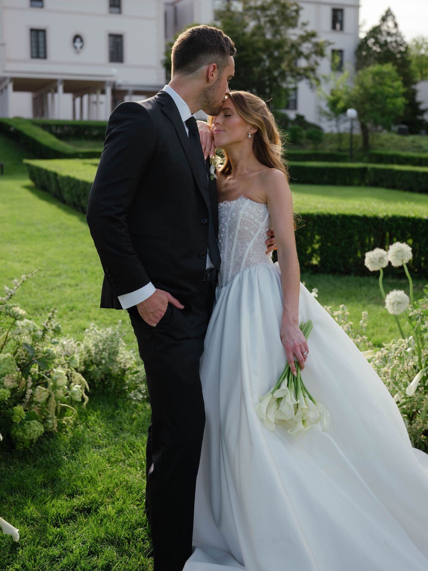 Couple shares a kiss in elegant garden, bride holding white calla lilies bouquet in light blue ball gown.