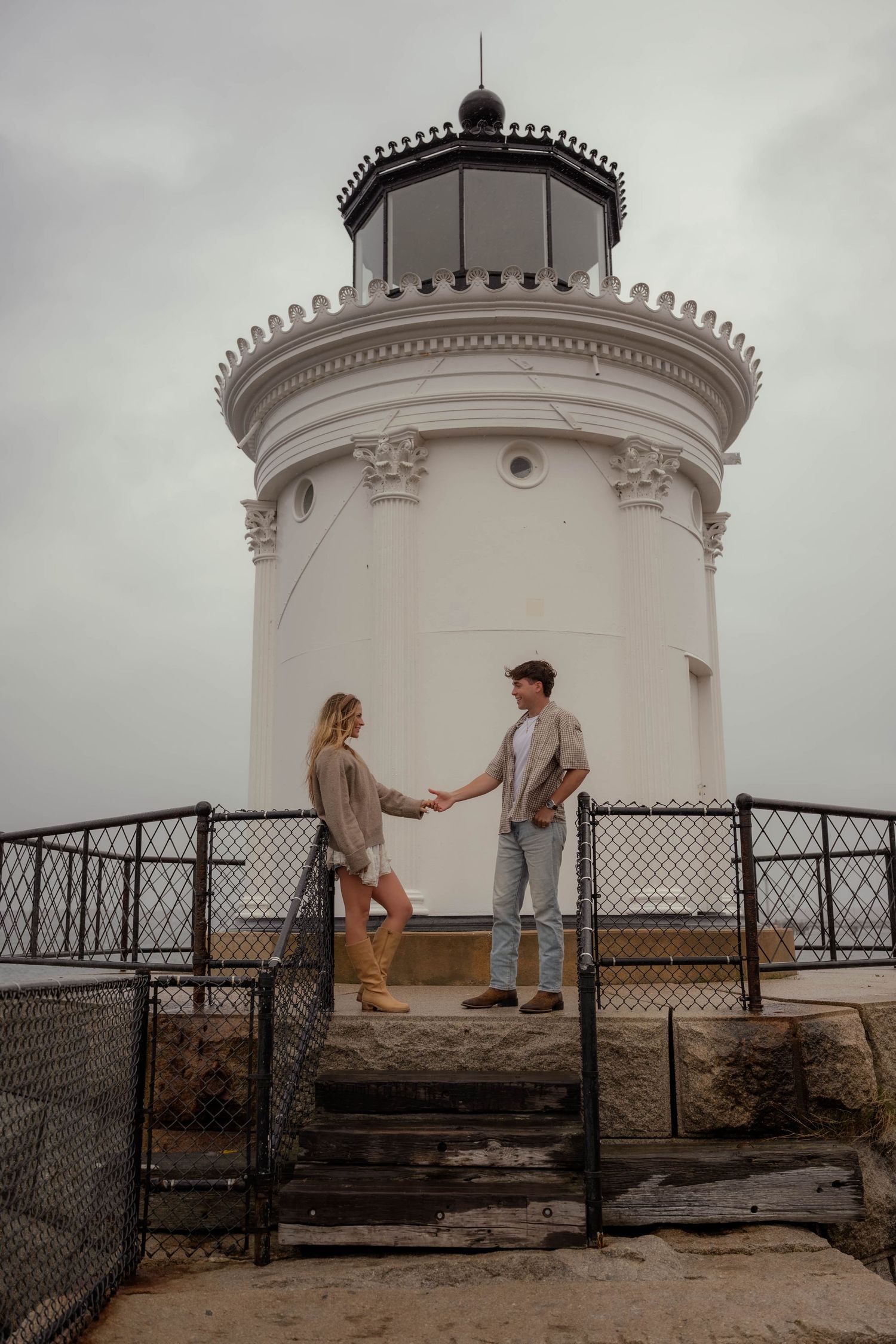 Young couple holding hands on stone steps in front of a white lighthouse on cloudy day. Maine Lighthouse