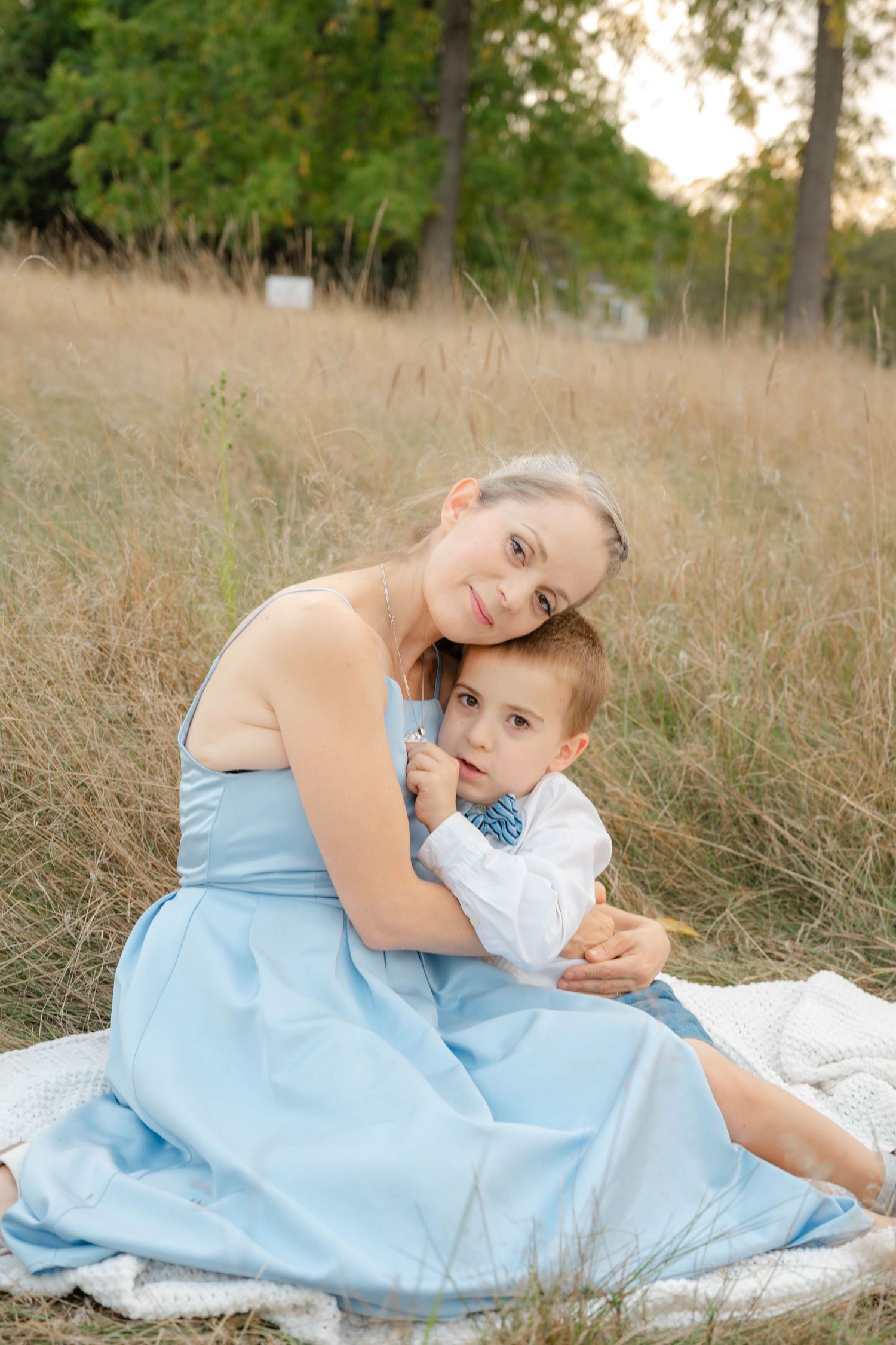 Young mother in blue dress sitting in golden field tenderly embracing her little boy in a white shirt with bow tie.