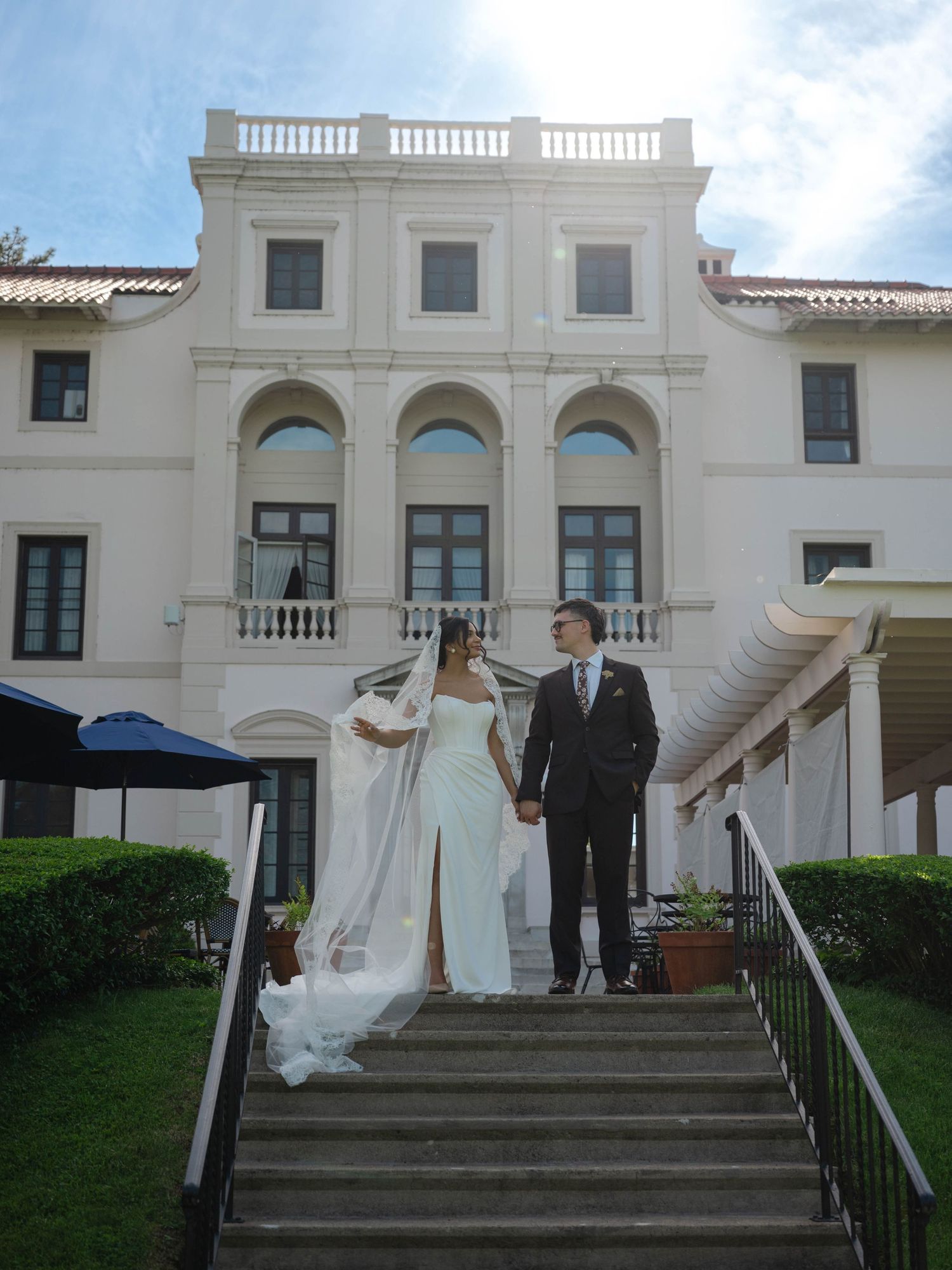 Bride and groom holding hands while walking down steps of a grand white neoclassical mansion on their wedding day. Central New York
