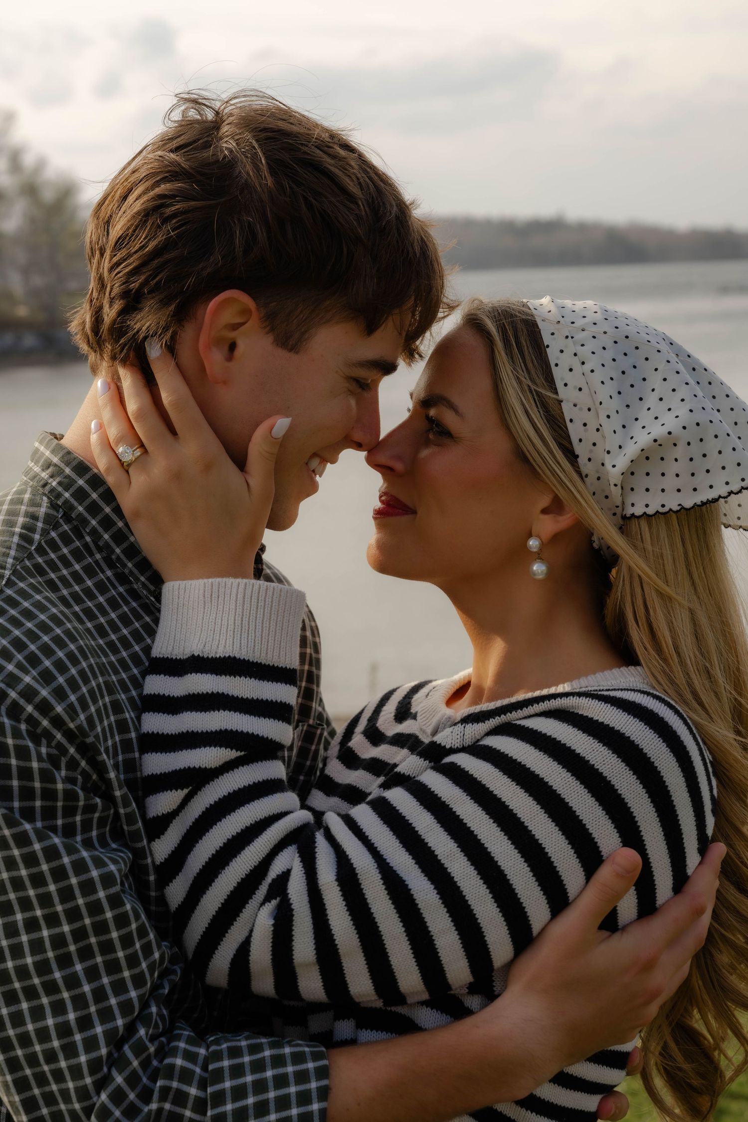 Couple sharing a romantic nose-to-nose moment outdoors near a lake, woman wearing striped top and polka dot headscarf.
