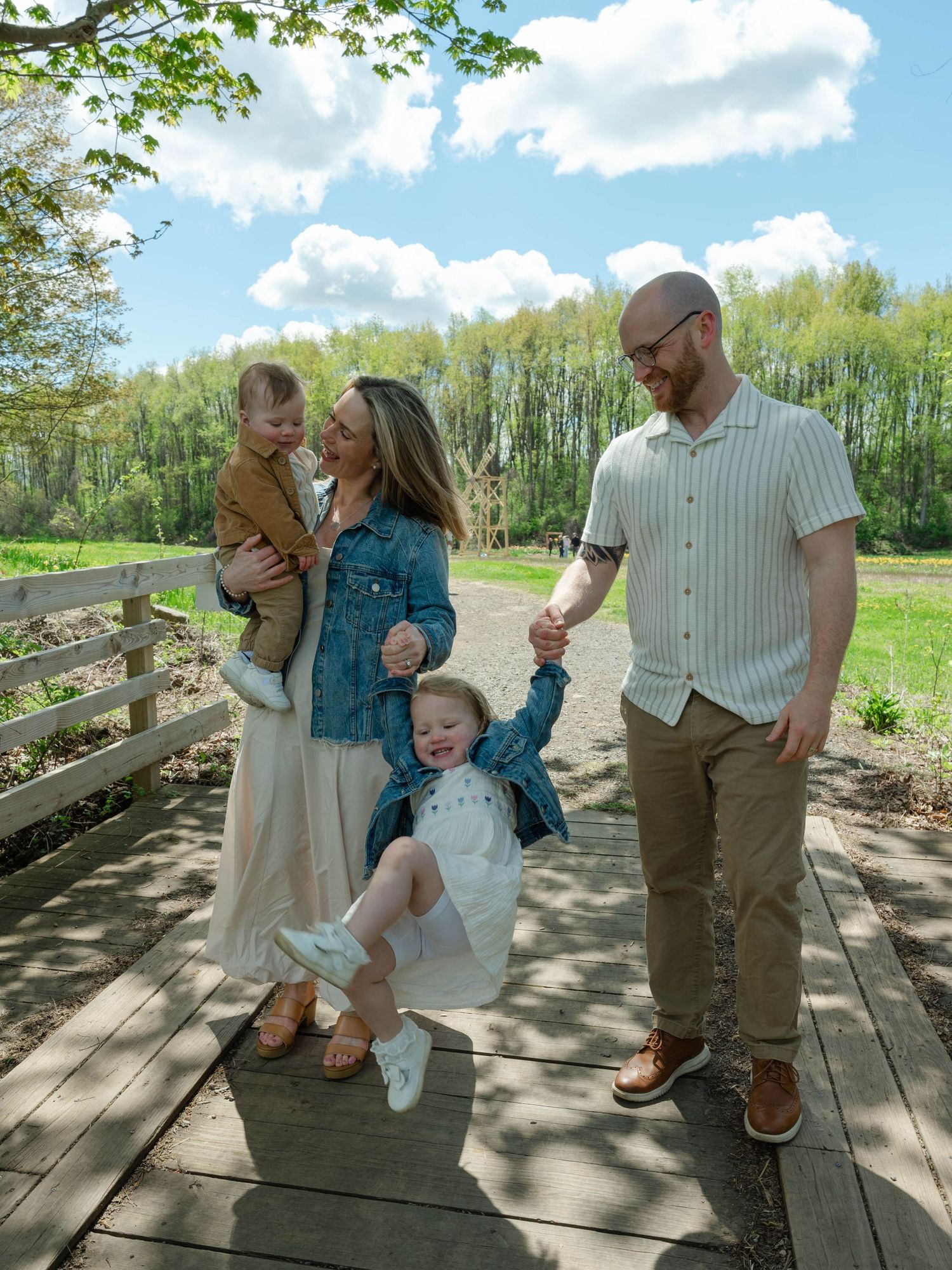 Joyful family of four playing on wooden bridge path with green trees and blue sky backdrop.