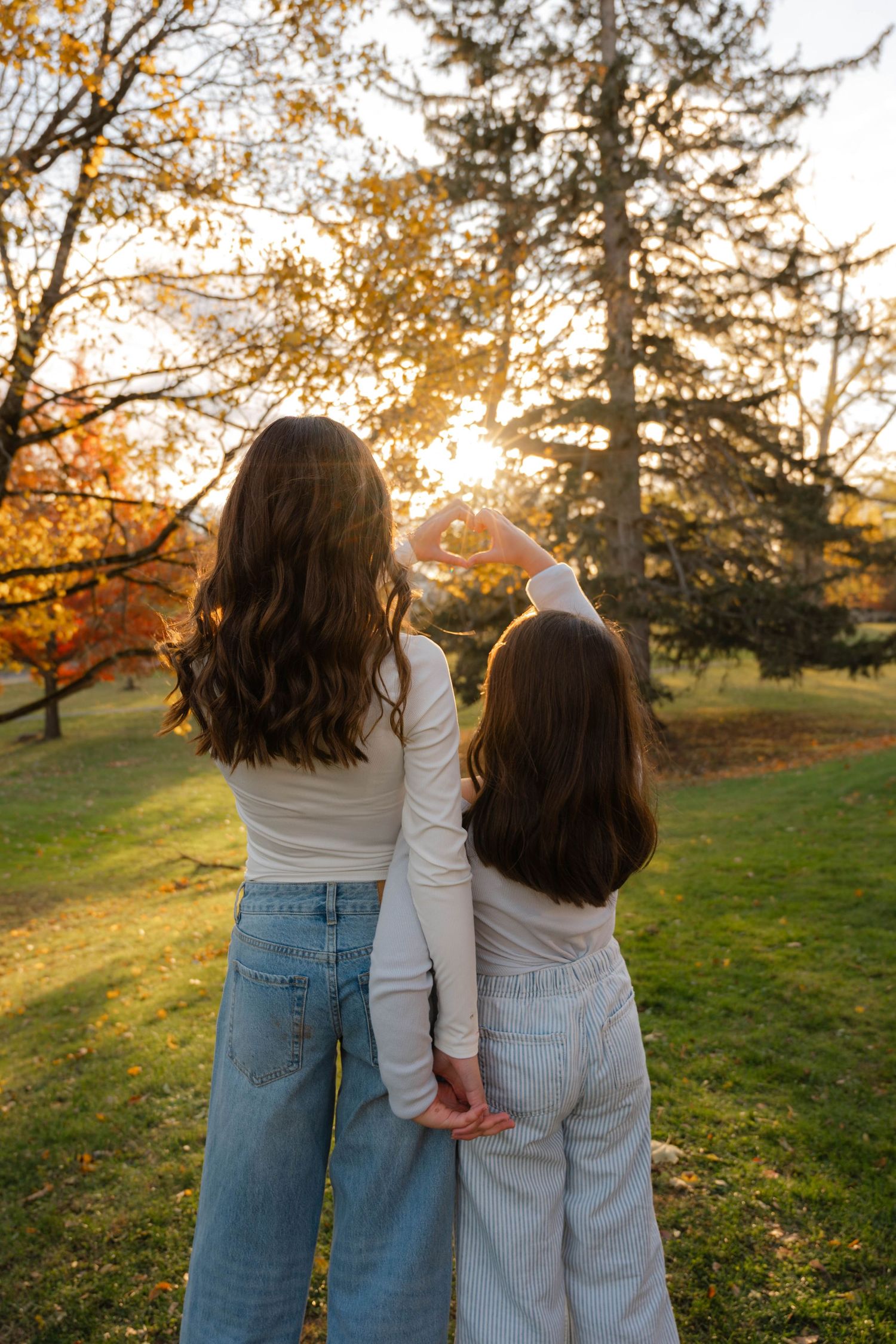 Two dark-haired girls in white tops stand together facing a glowing autumn sunset in a park.