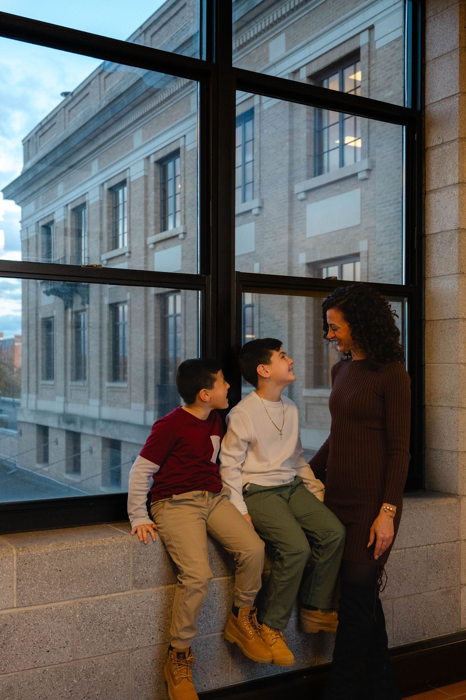 Pregnant woman in brown dress smiles at two boys sitting on window ledge in modern building.